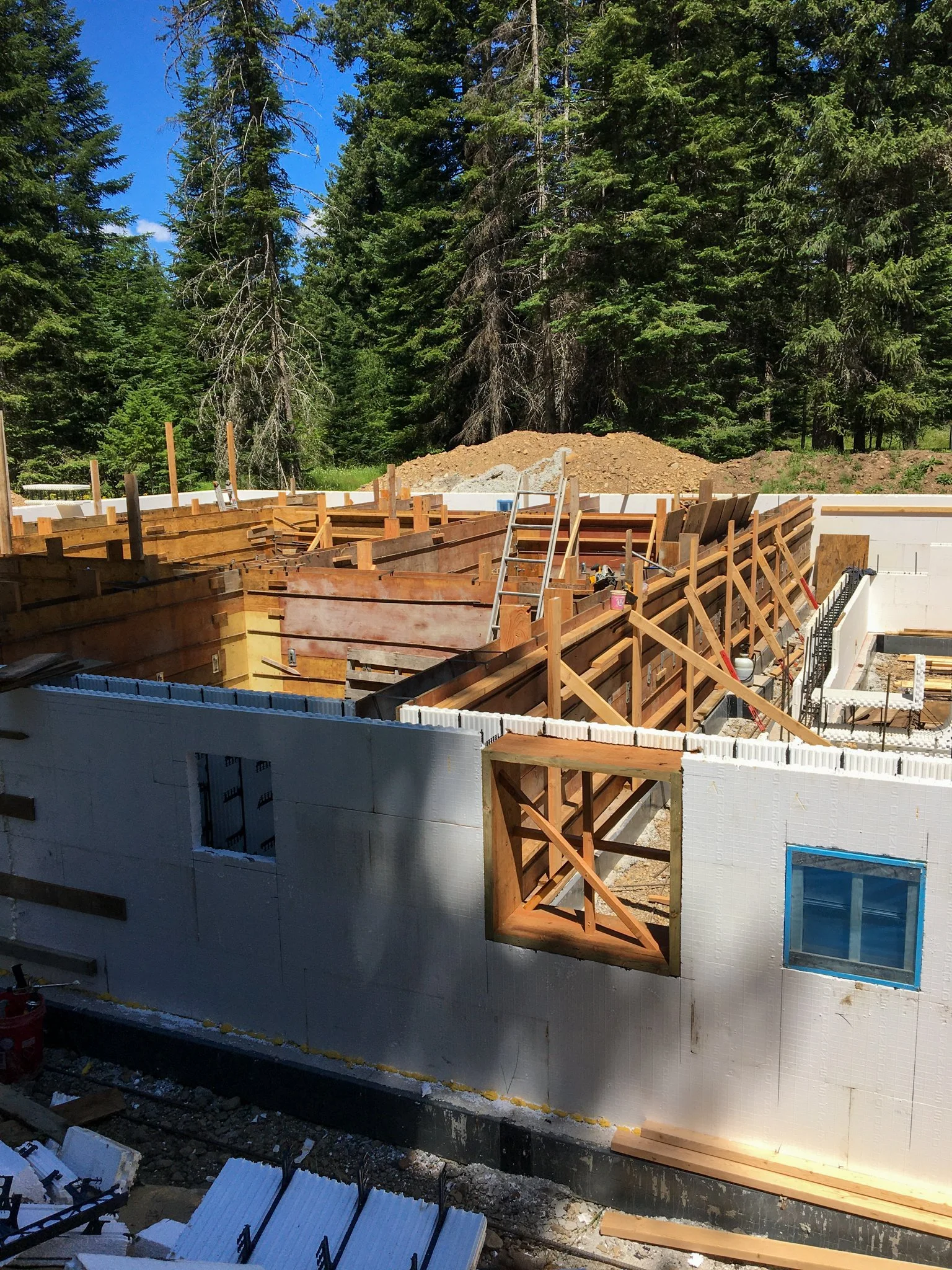 A house under construction with wooden framing and formwork, surrounded by tall forest trees on a sunny day.