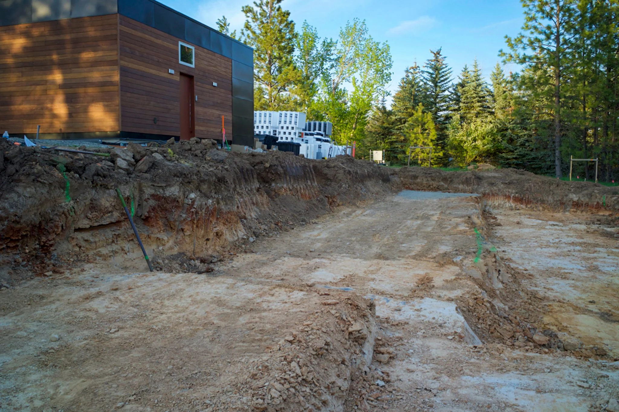 Construction site with excavated dirt in front of a modern house with wood siding and metal accents, surrounded by tall green trees under a clear blue sky.
