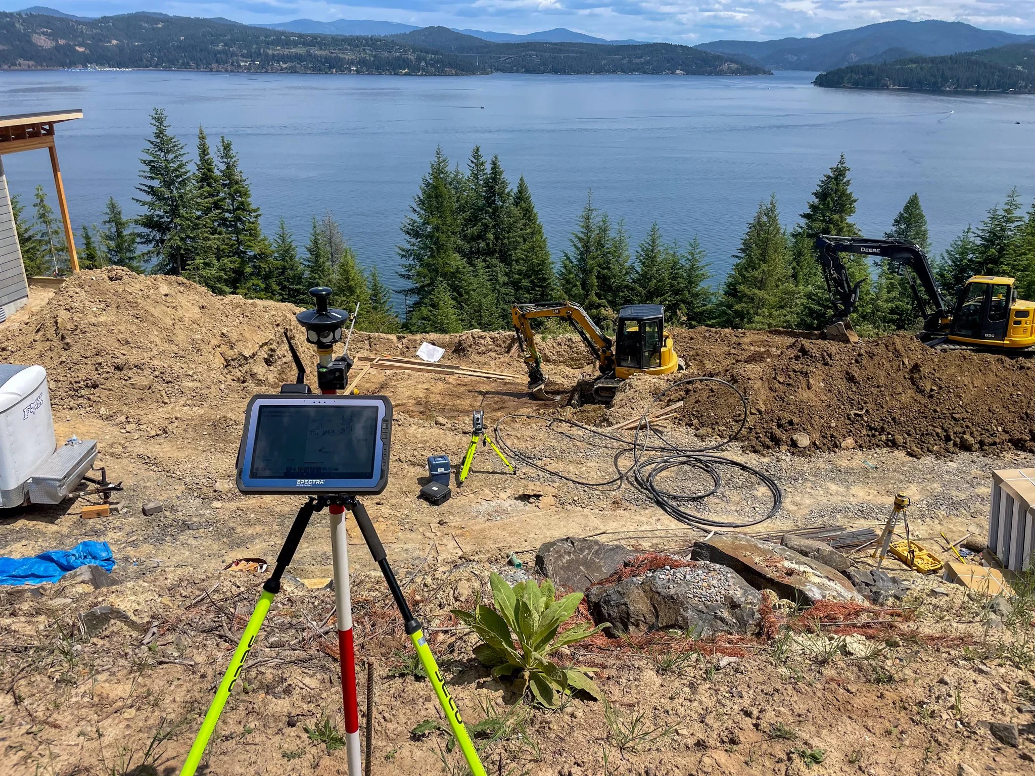 Construction site near a body of water with trees and mountains in the background. Several construction equipment and surveying tools are visible, including a tripod with measurement devices, a small excavator, and electronic equipment.