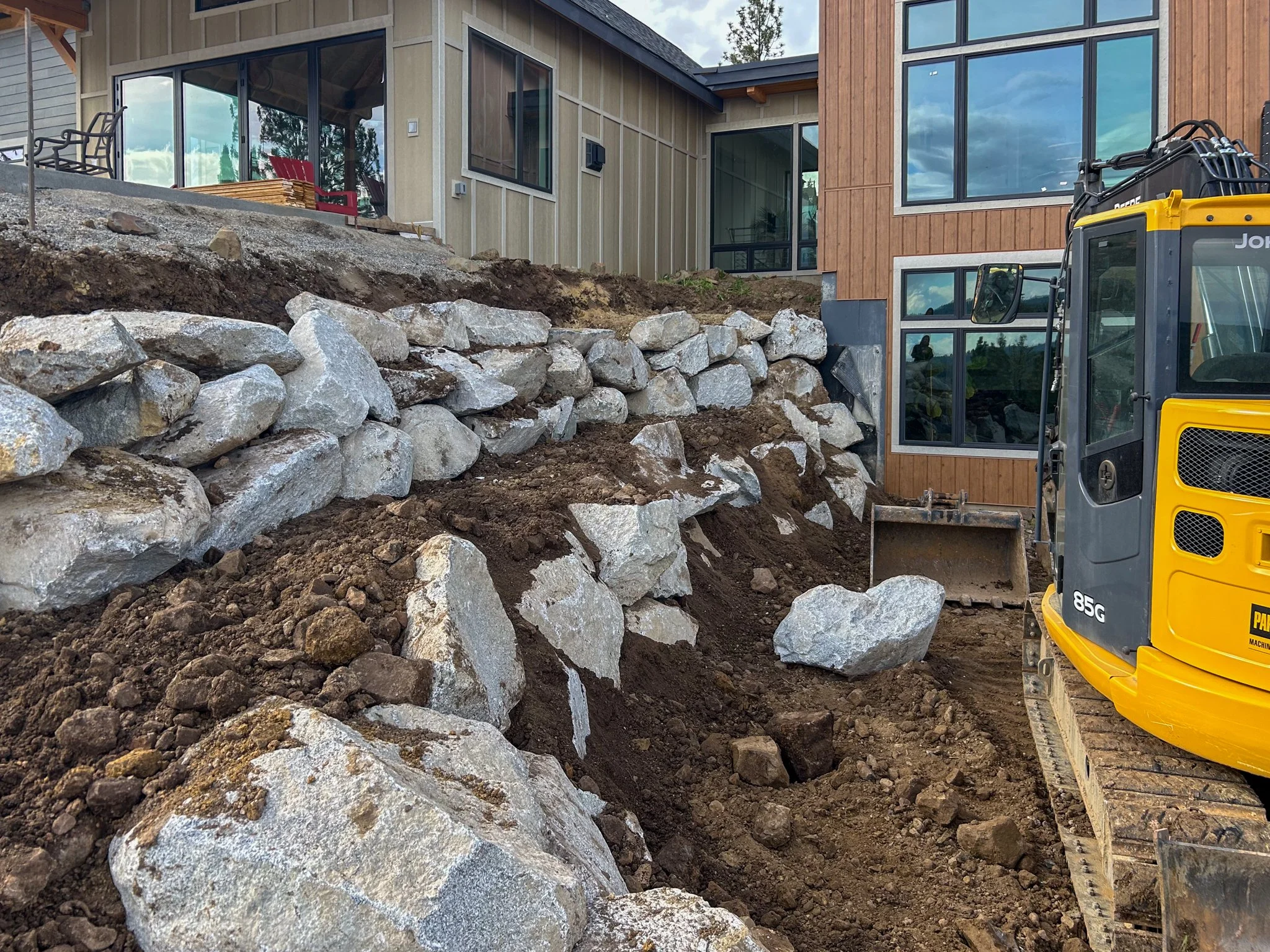 Construction site with large rocks and dirt in front of a modern house, yellow excavator in the foreground, wooden exterior, and large glass windows.