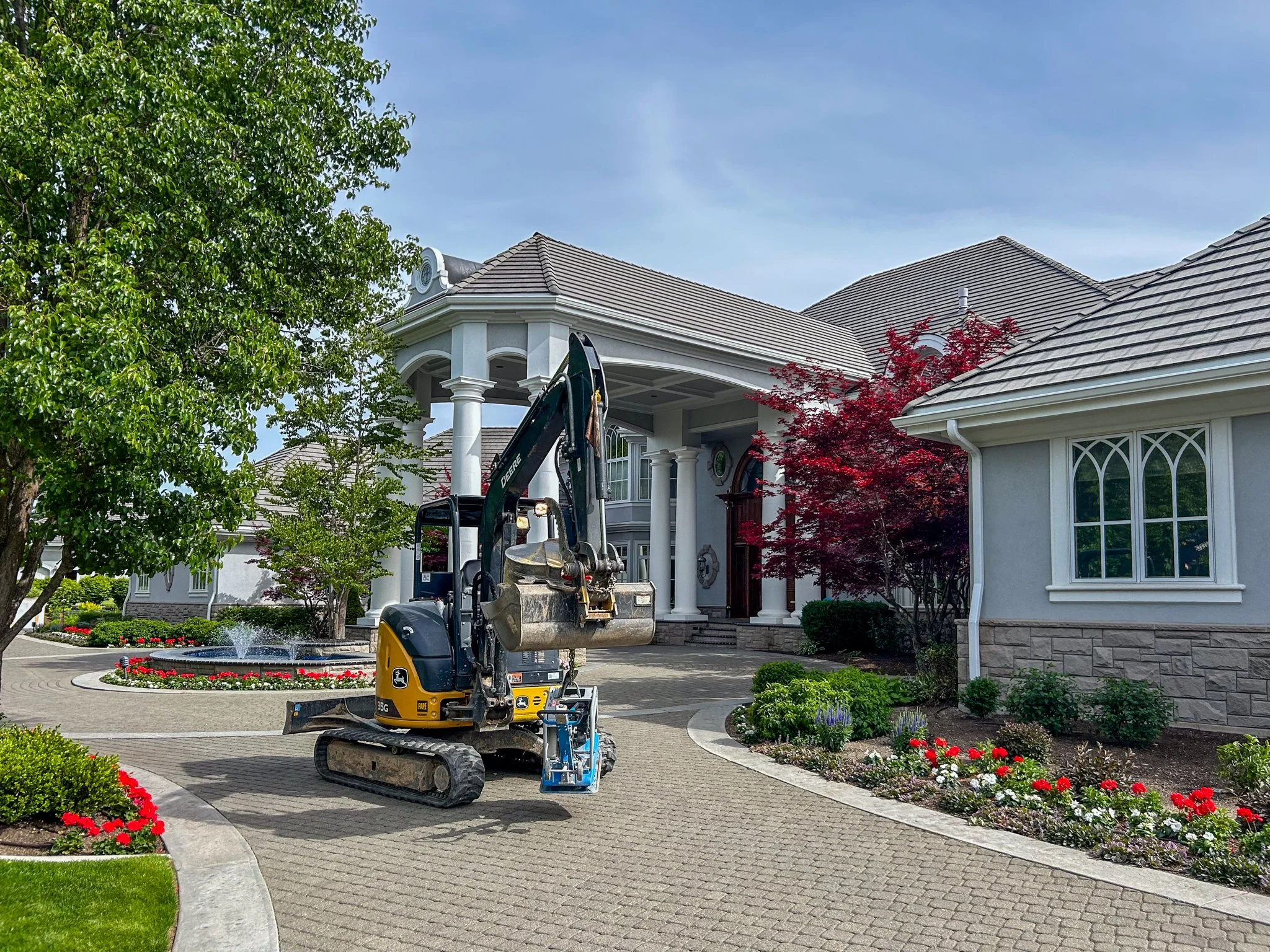 A two-story house with a large front porch, surrounded by a well-maintained garden with colorful flowers and trees. A mini excavator is parked on the driveway, likely for landscaping or construction work.