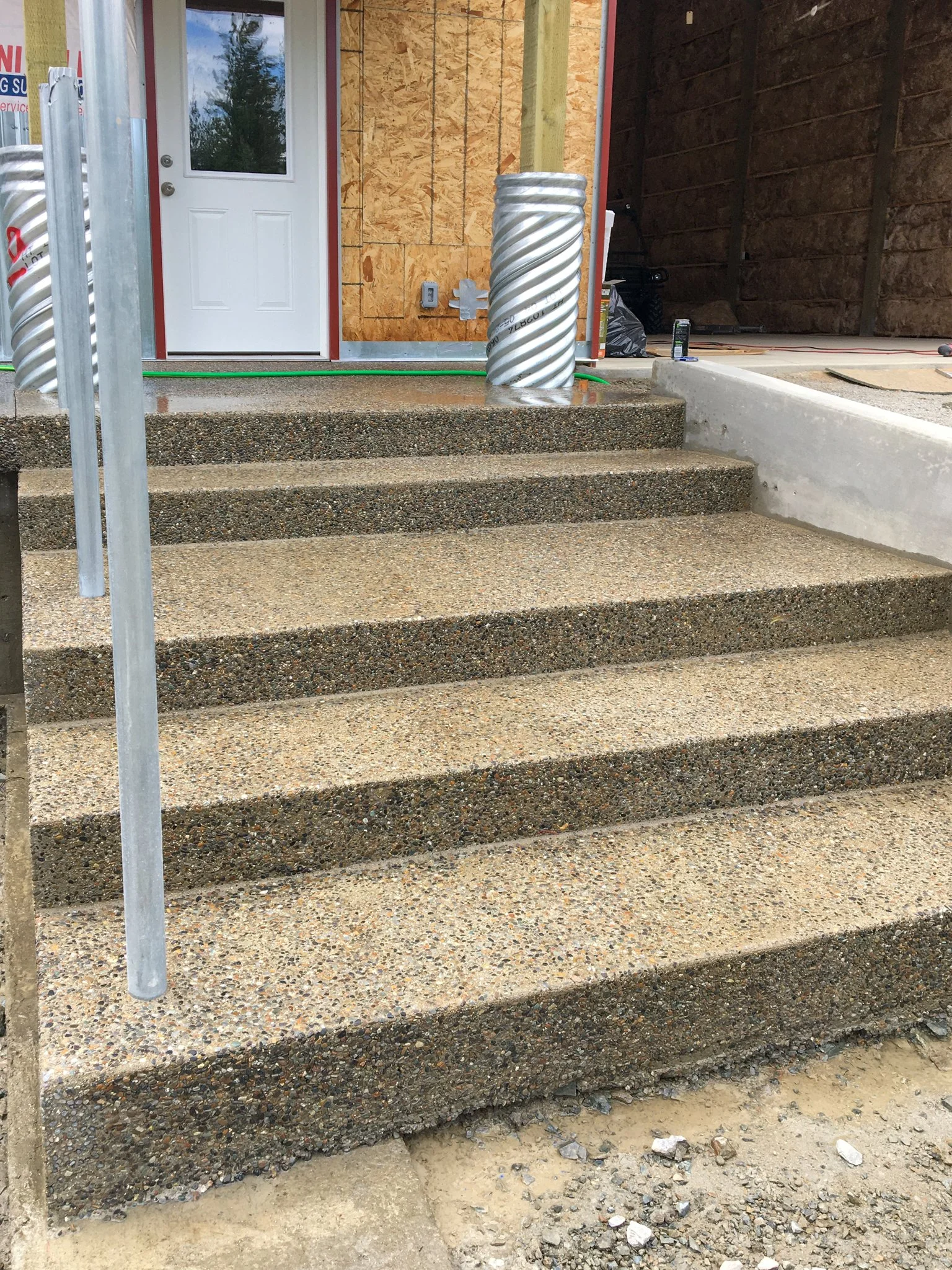 Construction site with gravel stairs leading to a door, with a protective metal railing on the left and construction materials on the right.