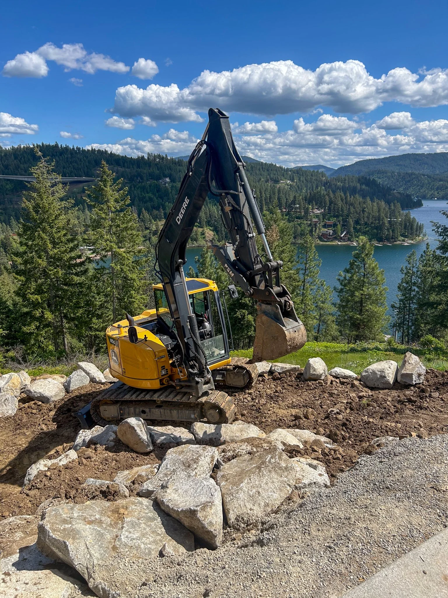 A yellow John Deere excavator working on a rocky hillside near a lake and forested mountains, with a partly cloudy blue sky overhead.