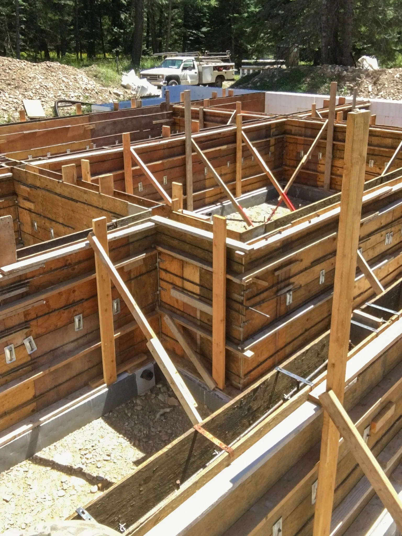 Construction site with wooden framing for a building, with support braces, a rebar framework, and a concrete foundation. A white pickup truck is in the background among trees.