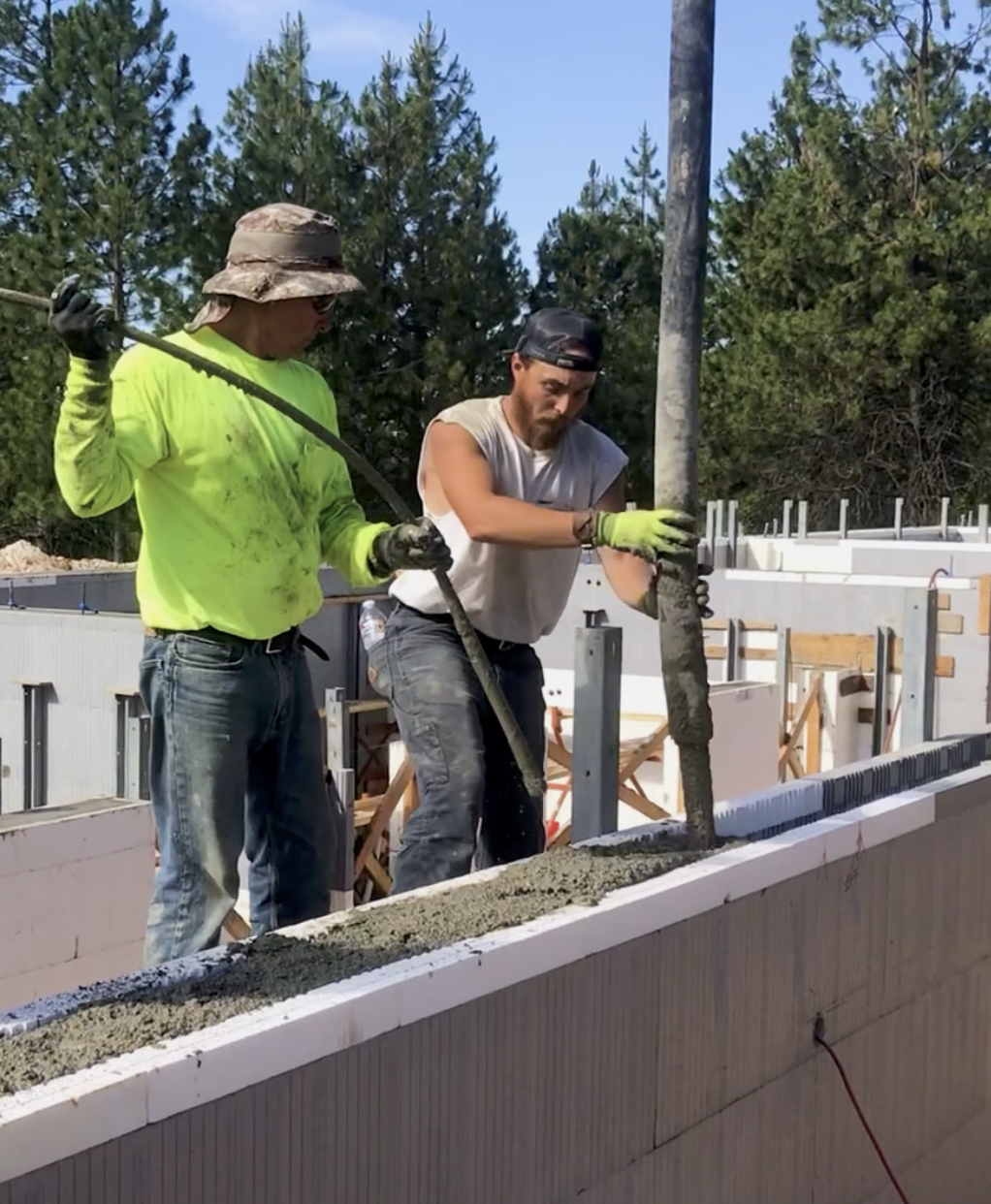 Two construction workers pouring concrete onto a building wall during construction, with trees in the background.