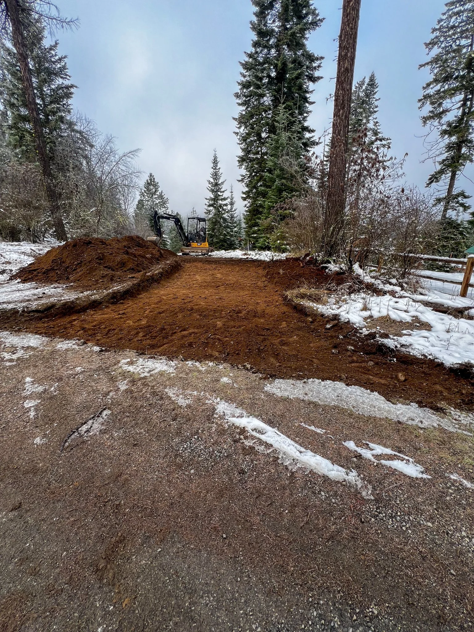 A dirt trail in a forest with tall pine trees and snow on the ground, a small yellow excavator in the background.