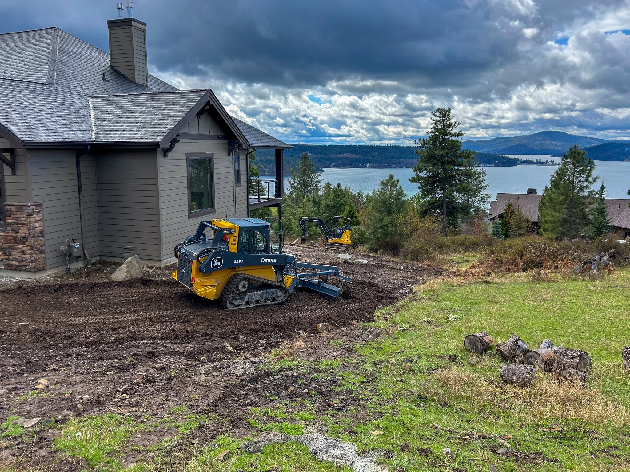 A house with gray siding and a shingled roof is under construction on a hillside overlooking a body of water and distant mountains. Construction equipment is visible in the foreground.