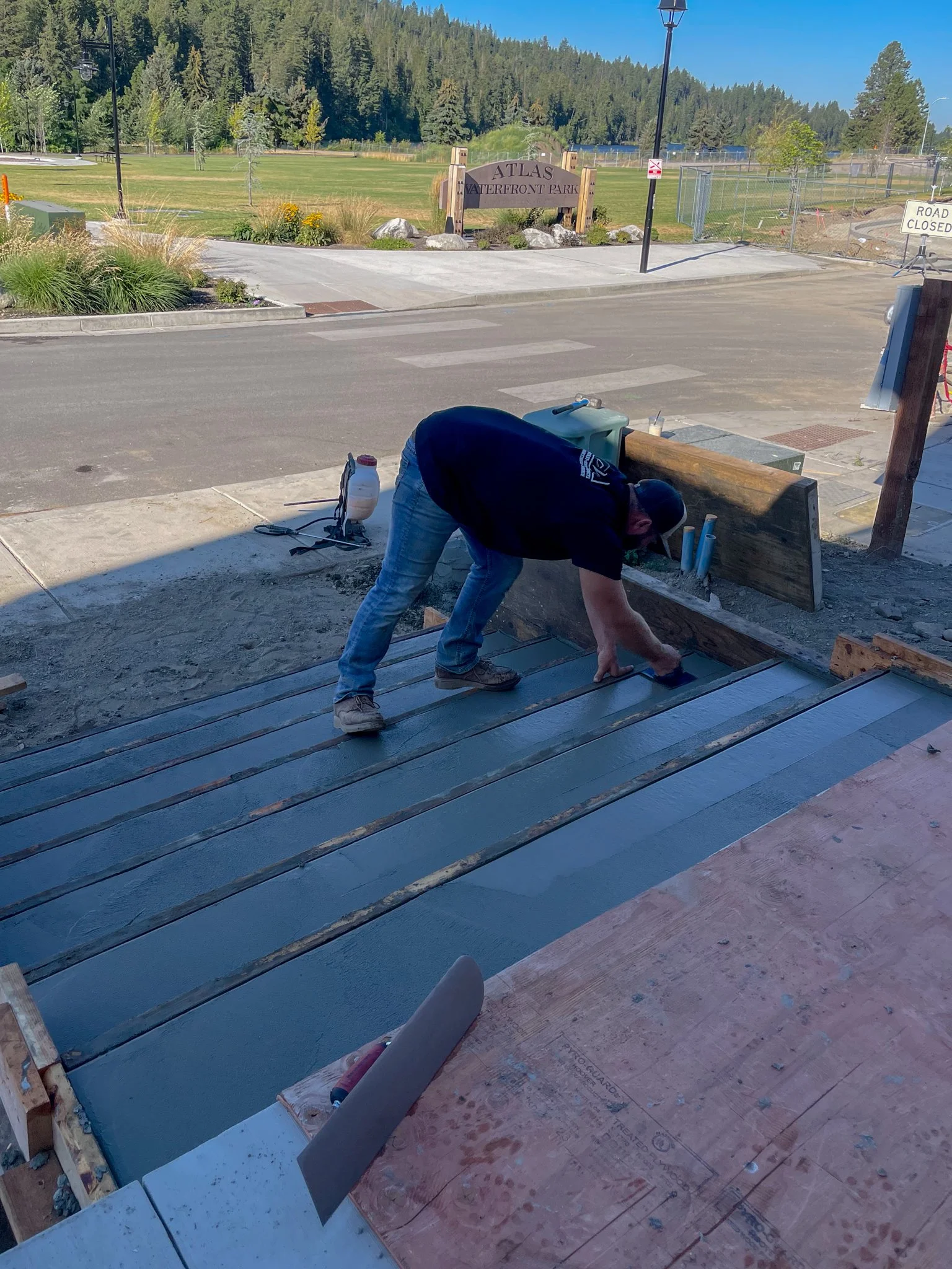 A construction worker bending over to work on a wooden deck or platform outside. The worker is wearing a cap, a dark shirt, and jeans. There are tools and construction materials on the ground nearby. In the background, there is a park entrance with a