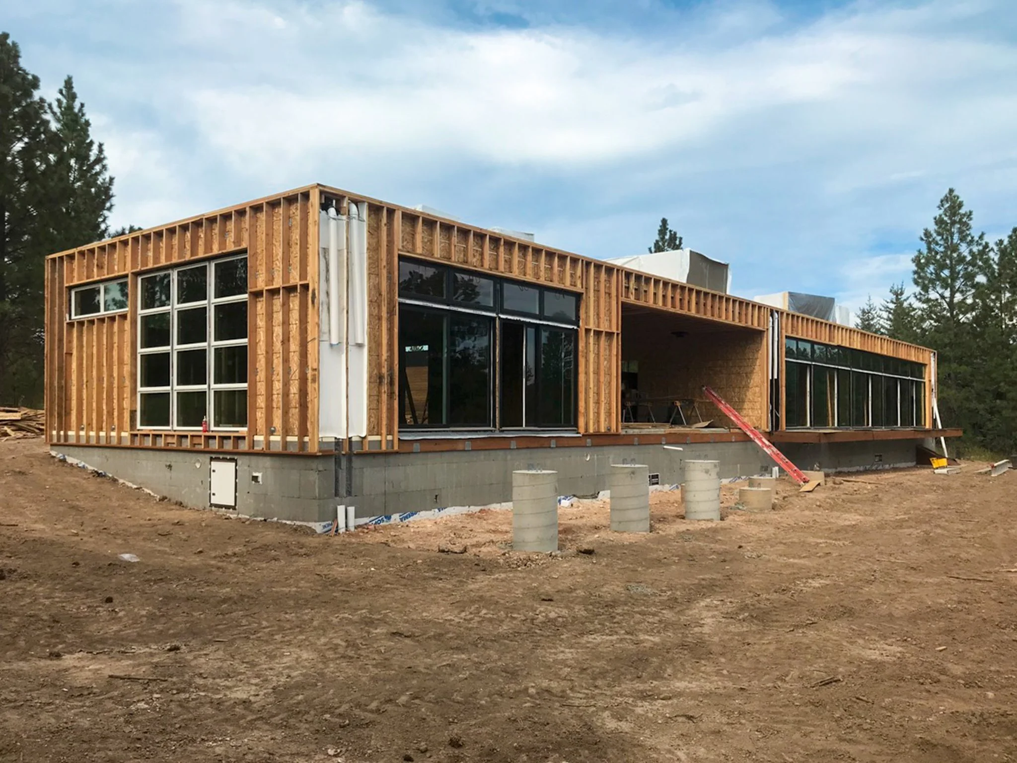A modern house under construction with a wooden frame, large glass windows, and construction materials on the ground, surrounded by tall trees and a cloudy sky.