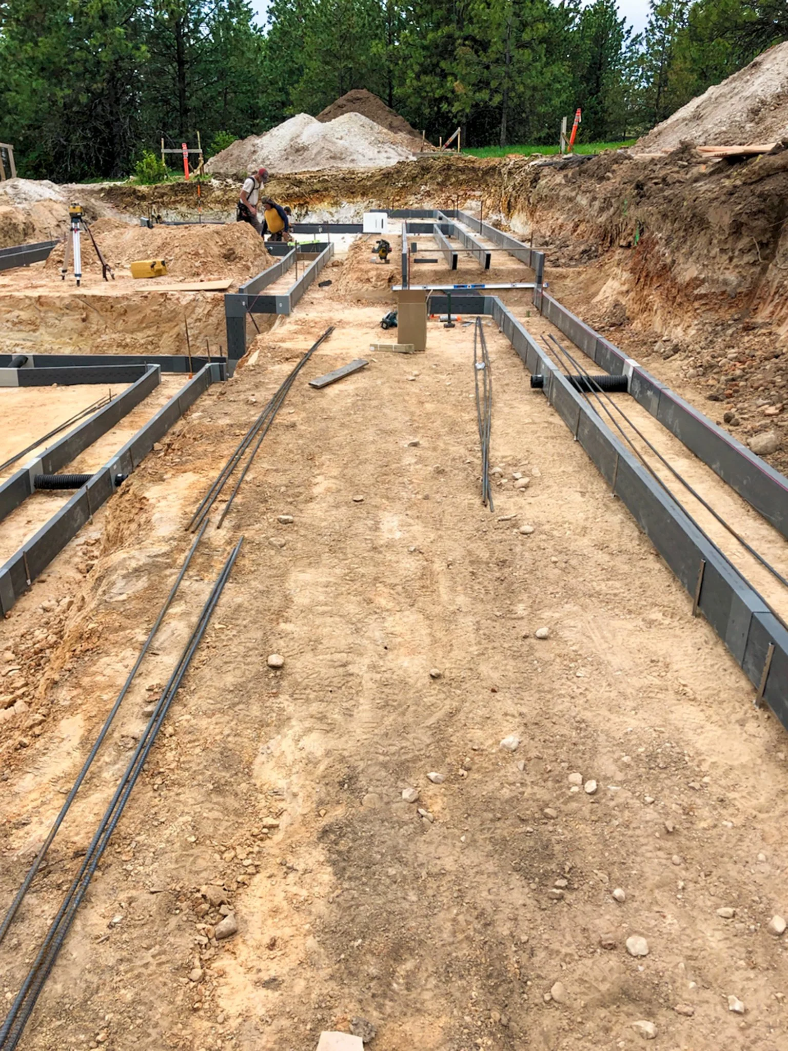 Construction site with foundation framework and workers preparing the site, surrounded by dug earth and trees in the background.