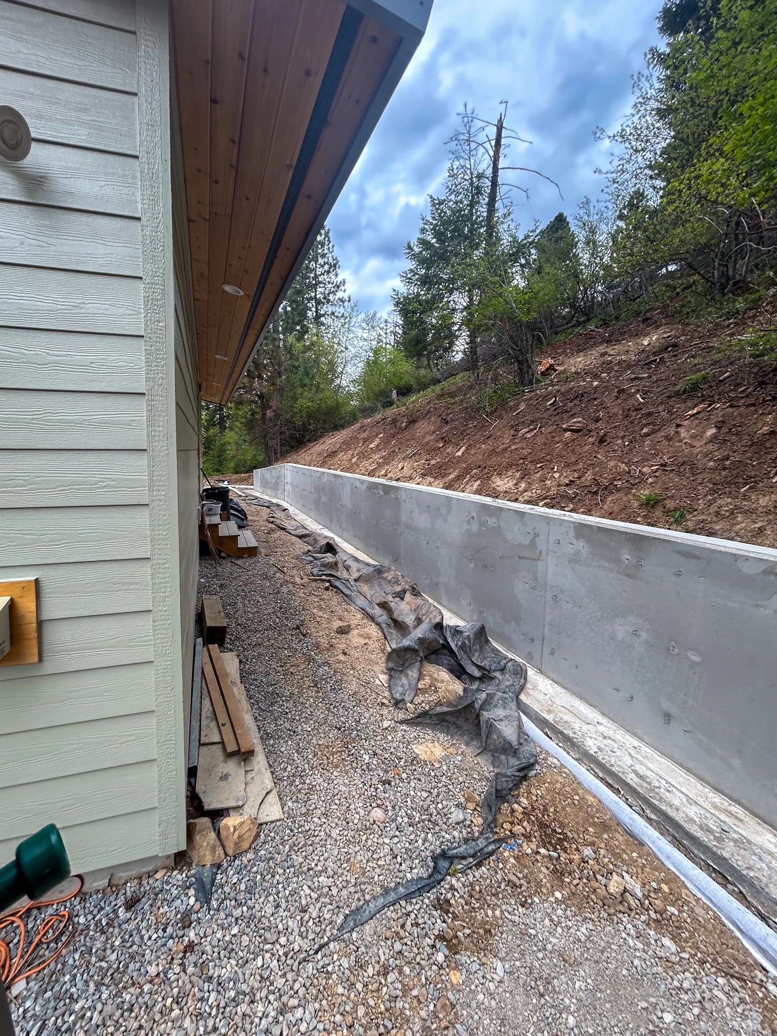 A construction site shows a gravel pathway next to a house with horizontal siding, with a wall on the left and a concrete foundation on the right. The house has a wooden overhang and some construction materials on the ground. Behind the house, there 