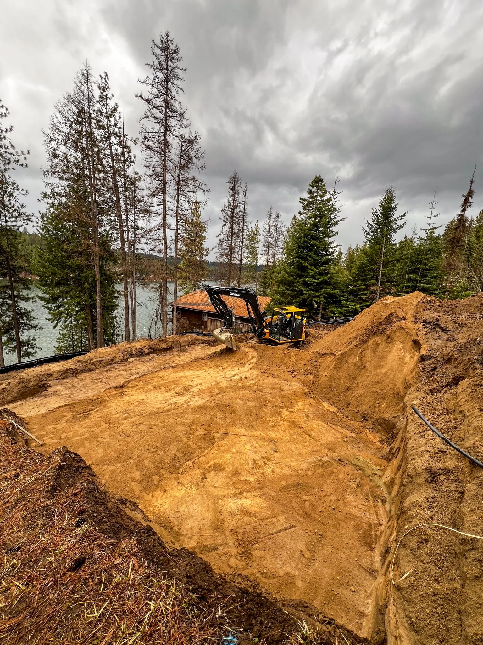 Excavation site with dirt mound, construction equipment, tall pine trees, and a lake in the background under gray cloudy sky.