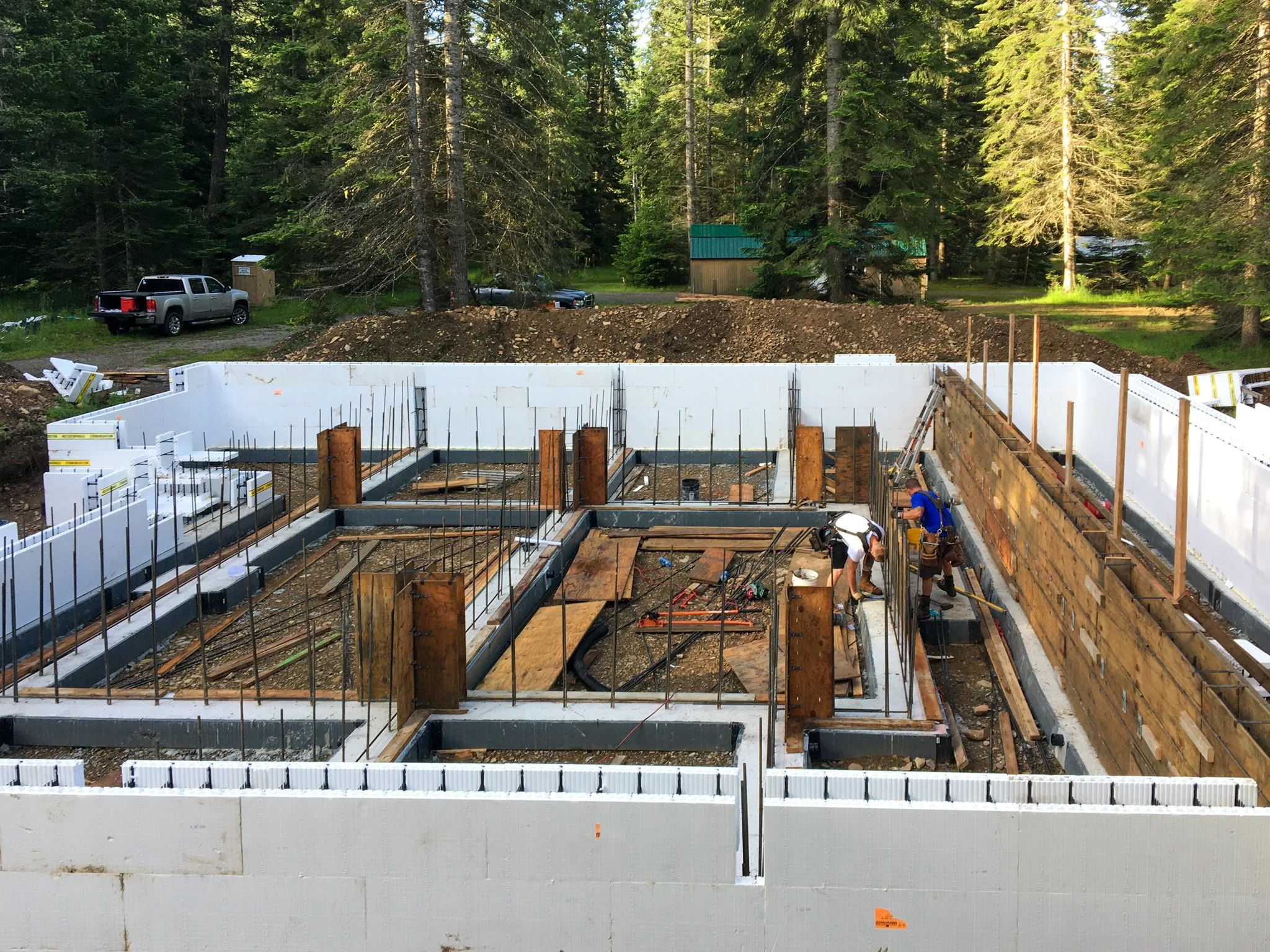Construction site with building foundation, workers, and surrounding trees in the background.