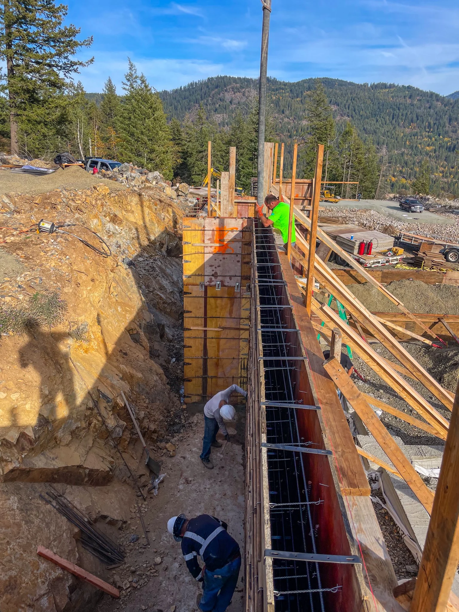 Workers constructing a concrete foundation on a mountainous construction site, with a wooden frame, rebar, and tools, surrounded by trees and blue sky.