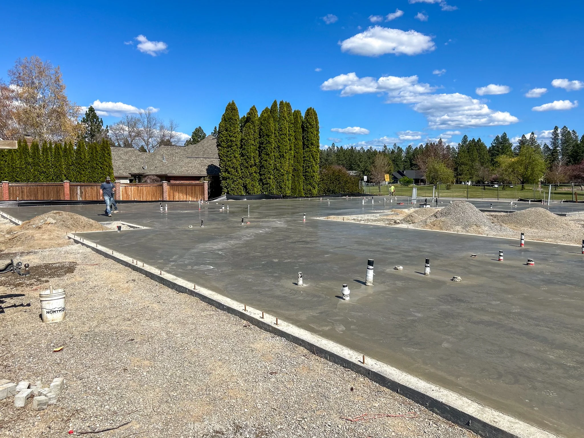 Construction site with freshly poured concrete slab, worker walking, and piles of gravel on the ground, with trees, houses, and a blue sky with clouds in the background.