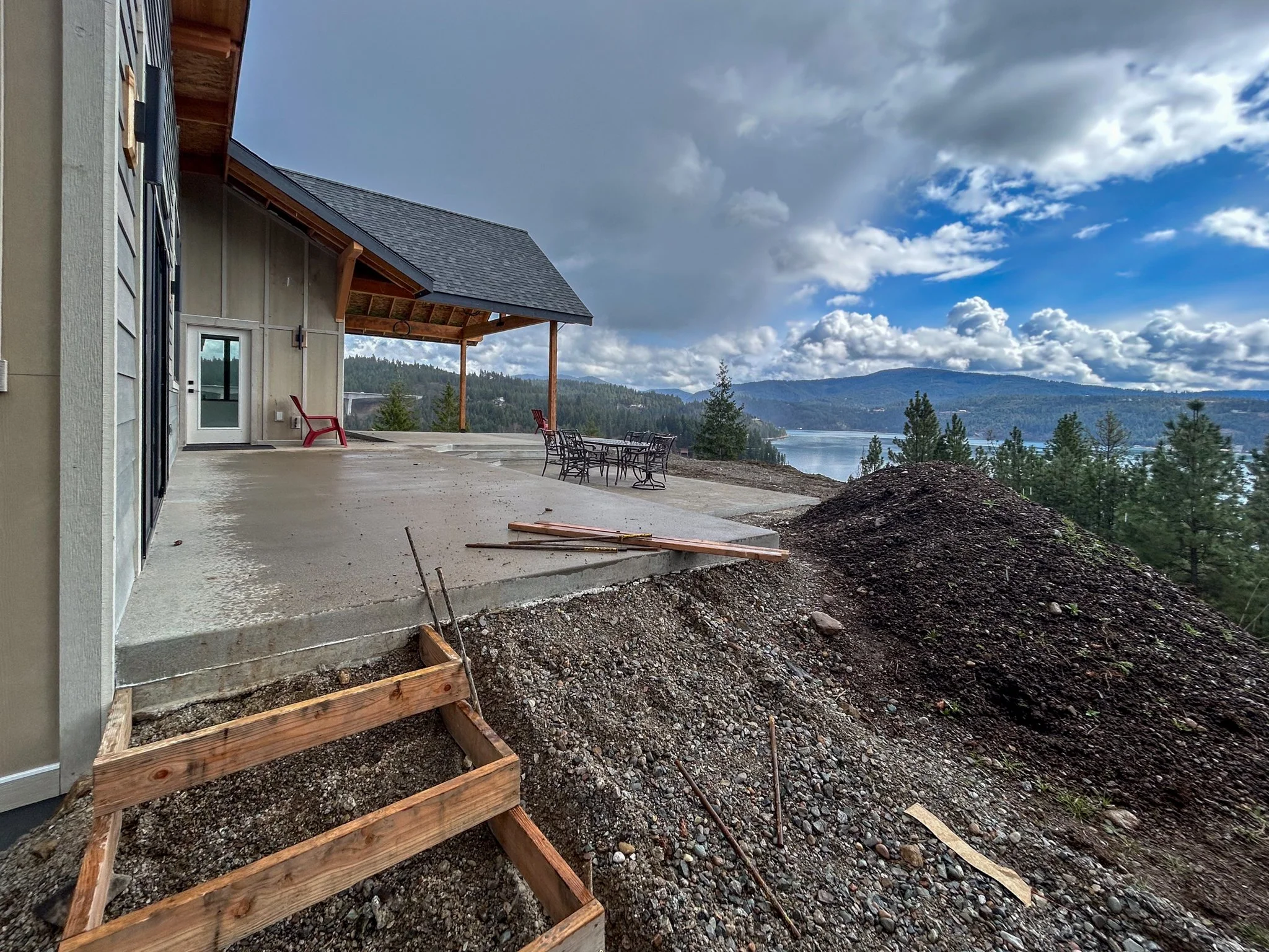 Construction site with an unfinished porch overlooking a lake and forested hills, cloudy sky.