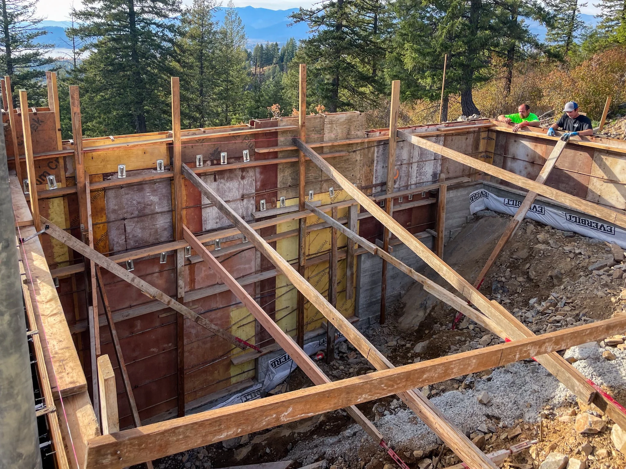 Construction site in a forest with wooden framework and two workers working on a concrete structure.