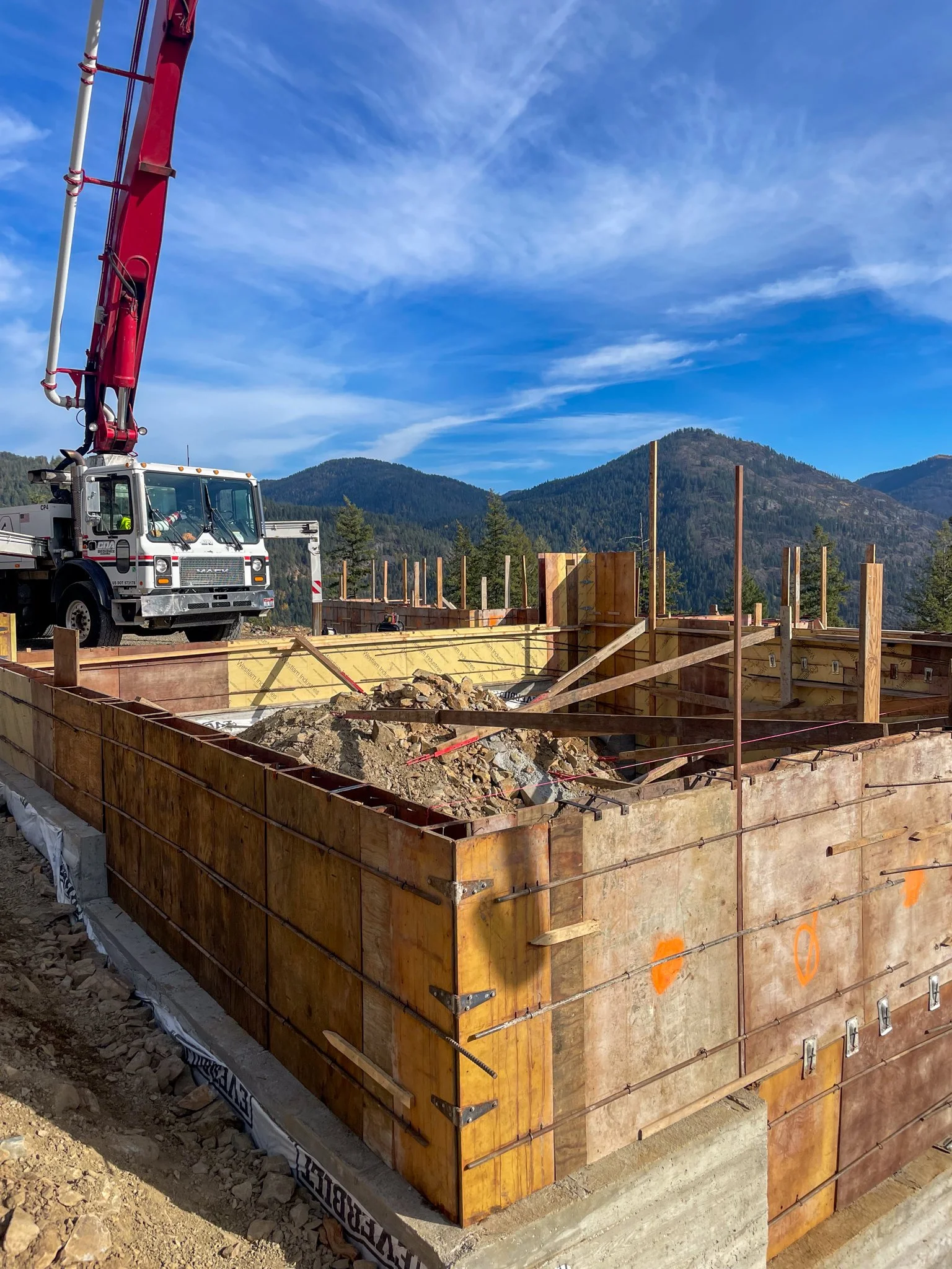 Construction site with a wooden framework, a crane, and scenic mountains in the background under a partly cloudy blue sky.