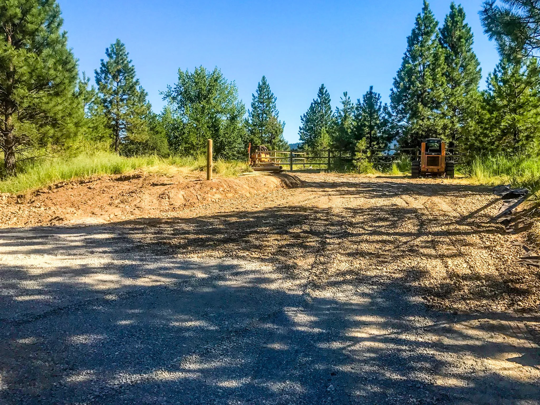 A dirt road leading to a gated area with construction equipment, surrounded by tall green pine trees under a clear blue sky.