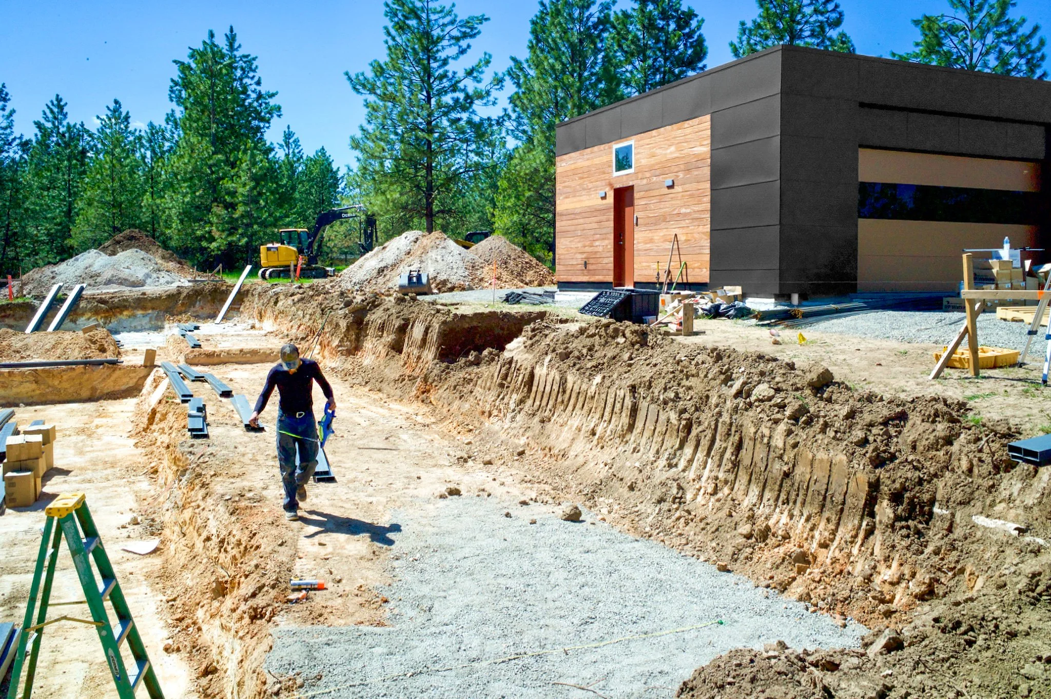 Construction site with excavation trenches, a worker walking, and a modern house with a wood and black exterior, surrounded by trees