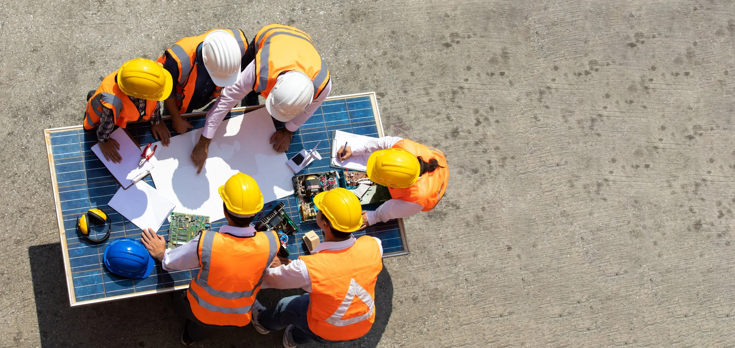 Top-down view of six construction workers wearing safety helmets and orange vests gathered around a solar panel on a concrete surface, working with tools and electronic equipment.