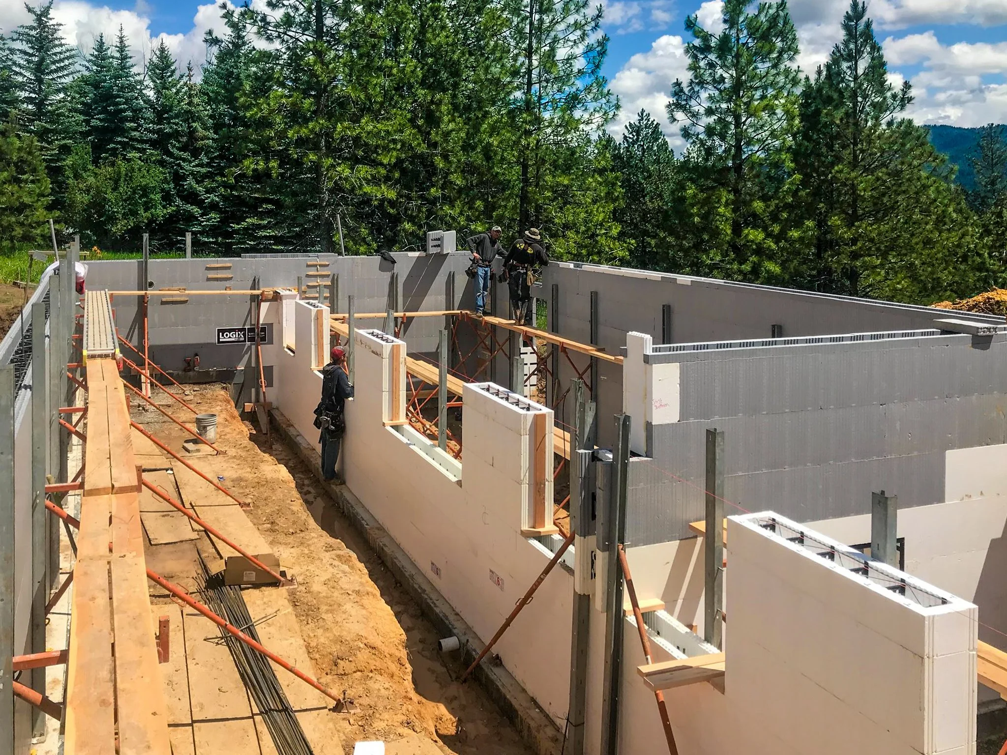 Construction workers building the walls of a house in a forested area with mountains in the background.