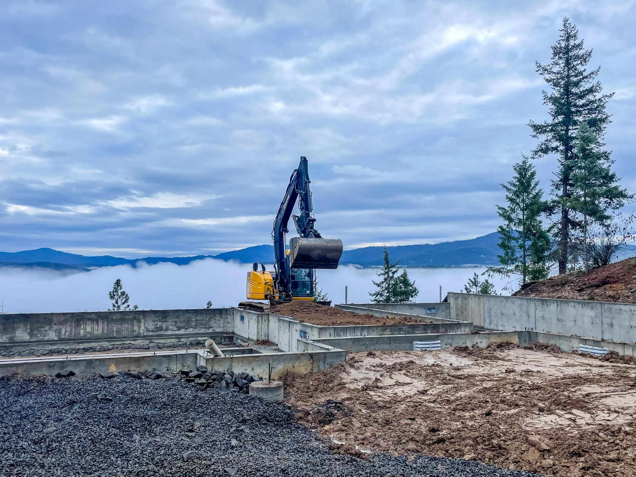 Construction site with a yellow excavator and concrete foundation in a mountainous area with trees and cloudy sky.