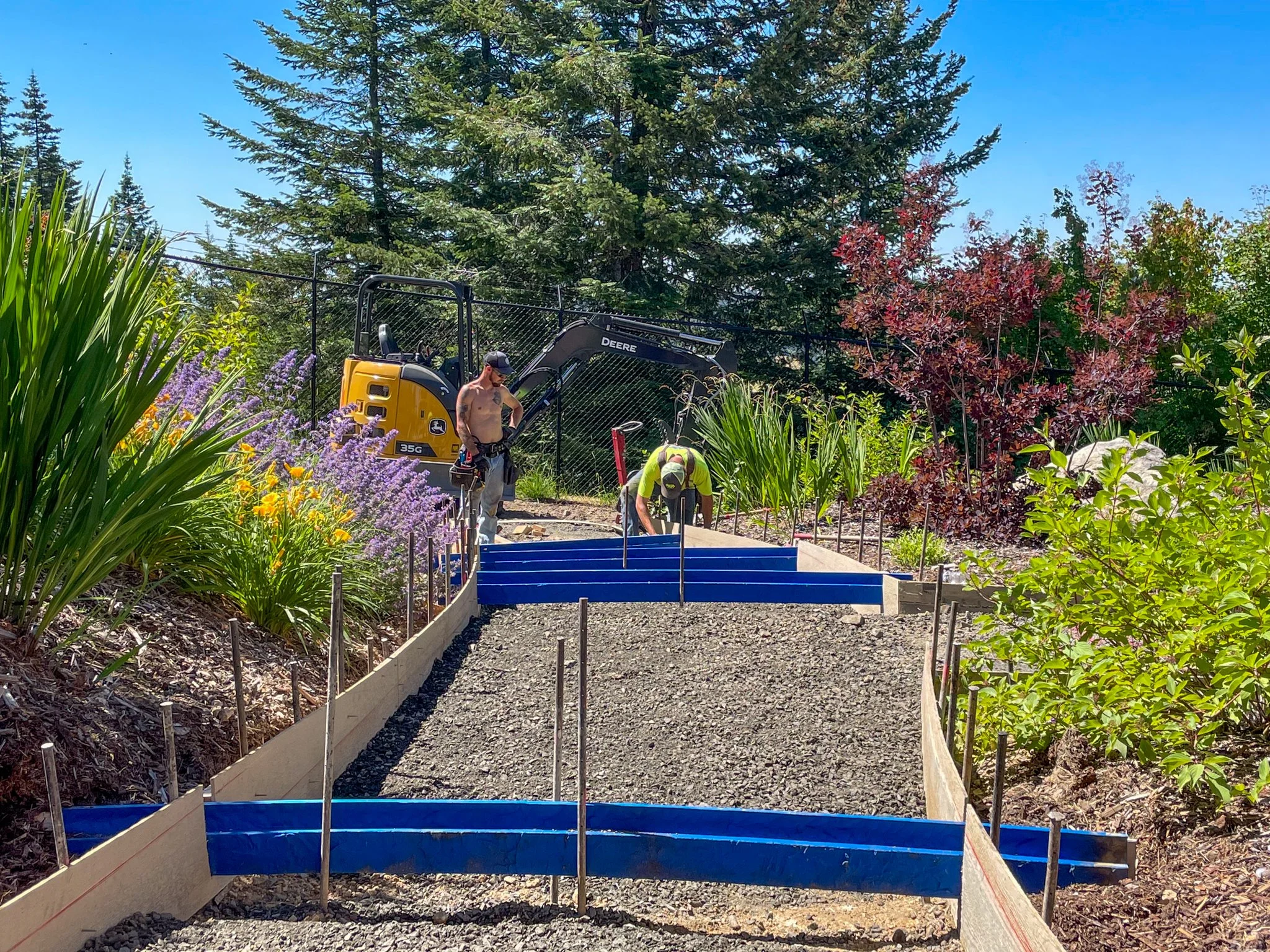A man standing on a gravel pathway in a garden, working on a construction project with a woman, with a yellow and black John Deere backhoe loader in the background, surrounded by green trees and colorful plants.