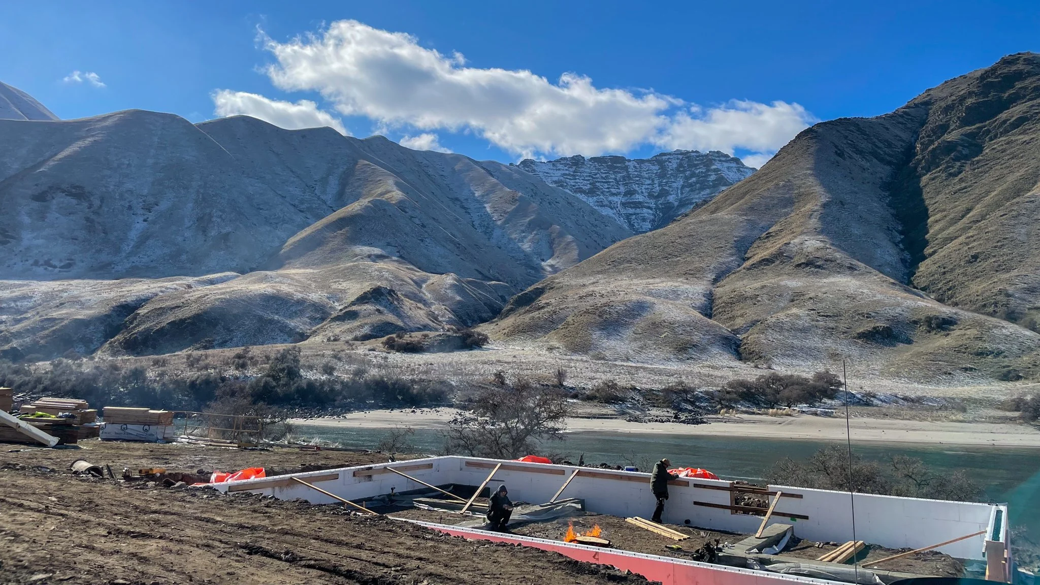 Construction workers building a boat along a riverbank with mountains in the background under a clear blue sky.