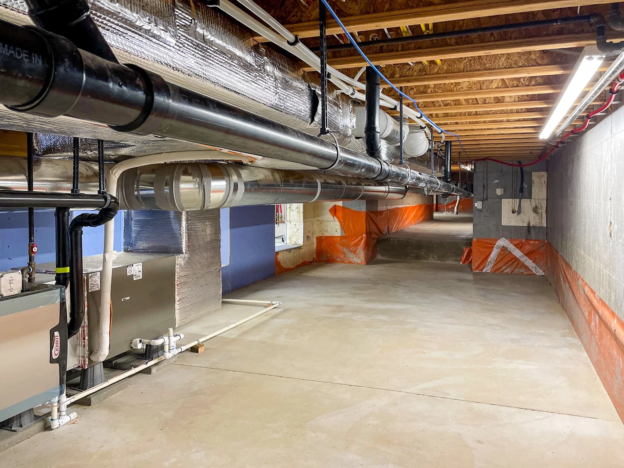 A basement with exposed wooden ceiling beams and concrete floor, featuring large pipes, electrical wiring, and construction debris covered with orange plastic, indicating ongoing electrical and plumbing work.