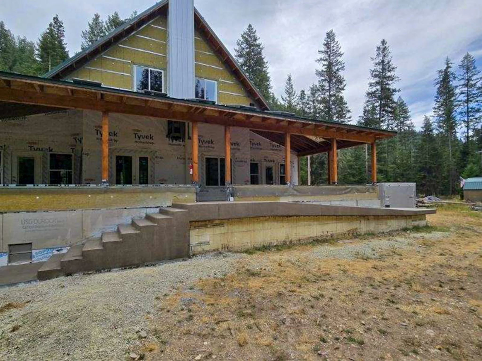 A house under construction with a steep gable roof and large windows, surrounded by a wooded area with tall trees. The house has unfinished exterior walls and a large covered porch supported by wooden beams.