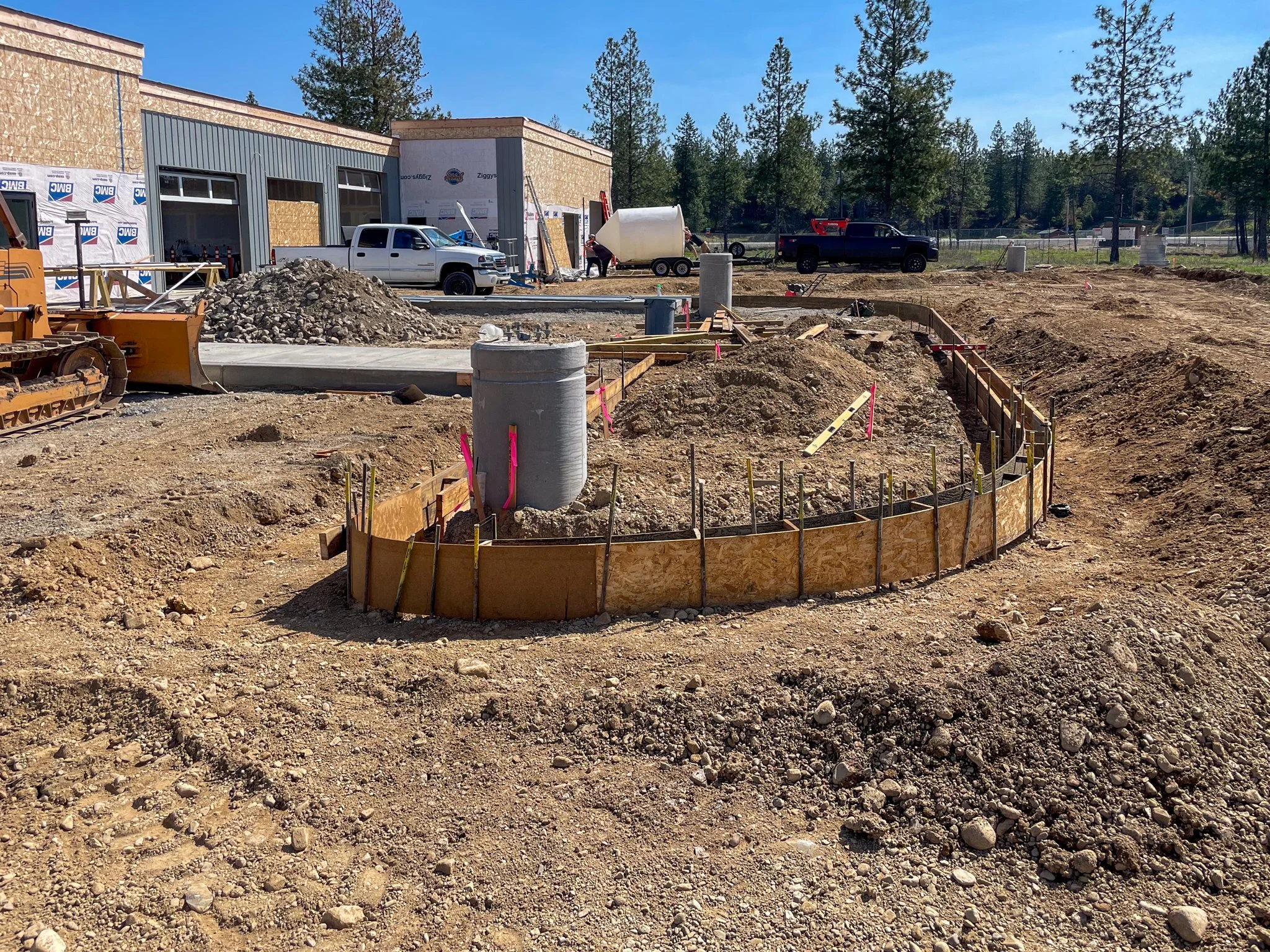 Construction site with partially built building, construction vehicles, and workers. Foreground shows excavated dirt with wooden forms for concrete. Background has trees and clear blue sky.