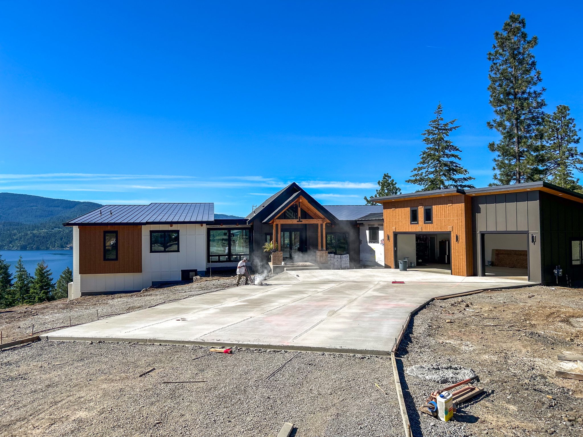 Under construction house with a concrete driveway and a lake view in the background, surrounded by tall trees, with building materials and workers present.