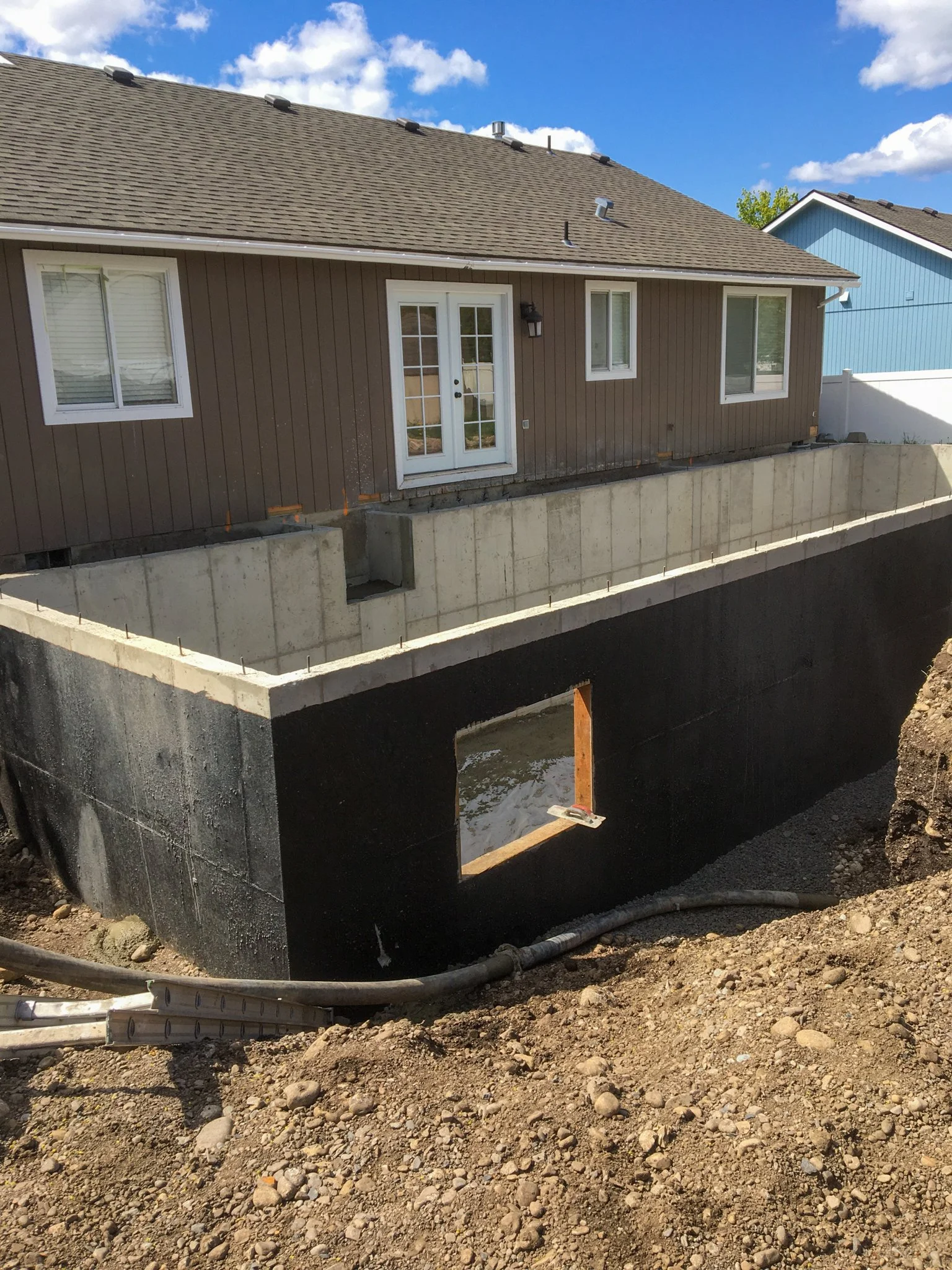 Construction site showing the foundation and basement of a house, with dirt and rocks around it. The house has brown siding, white door, and windows. Part of the roof and neighboring blue house are visible.