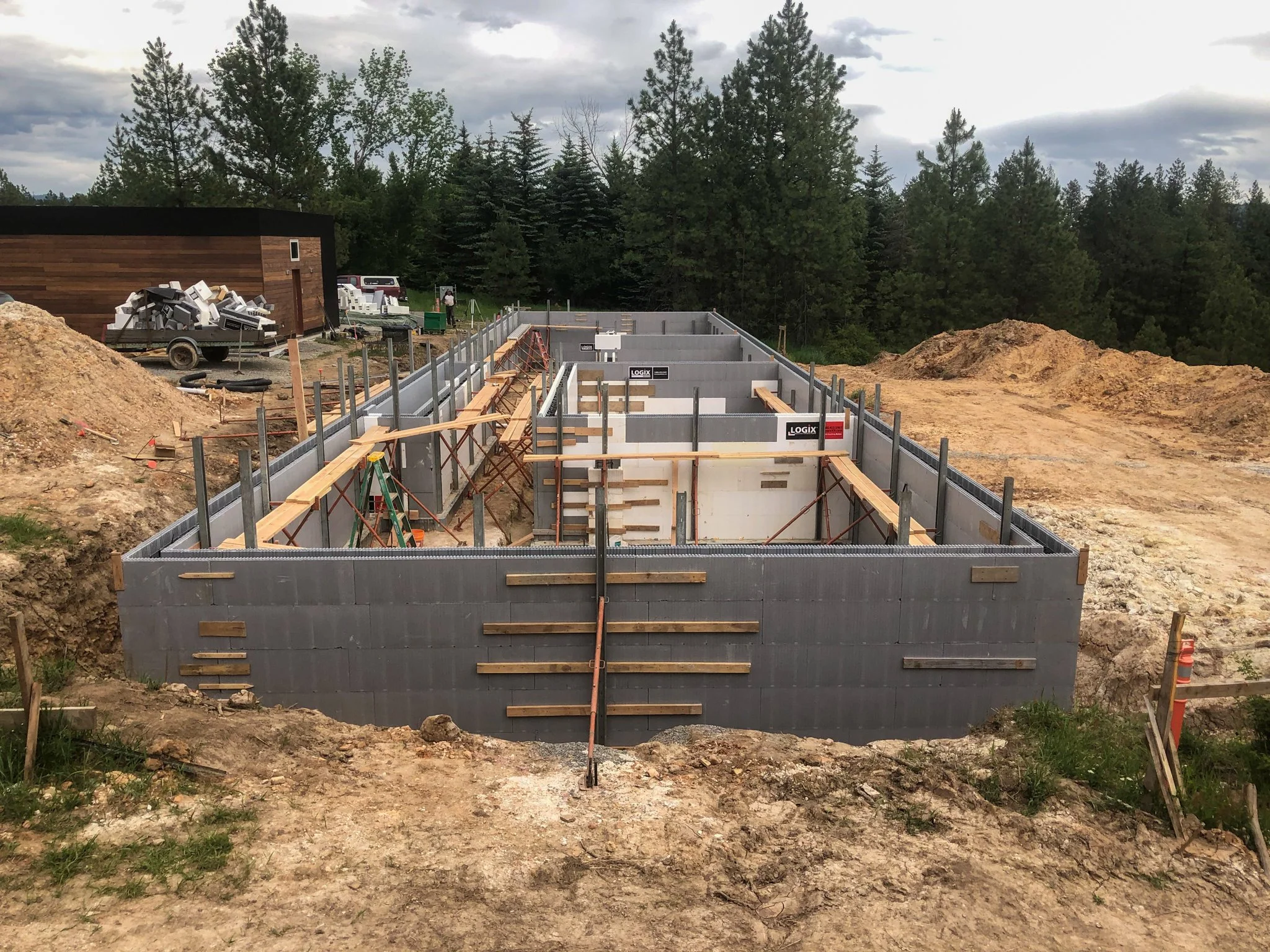 Construction site showing the foundation of a building being built with gray concrete blocks and wooden supports, surrounded by dirt and construction materials, with trees and an overcast sky in the background.