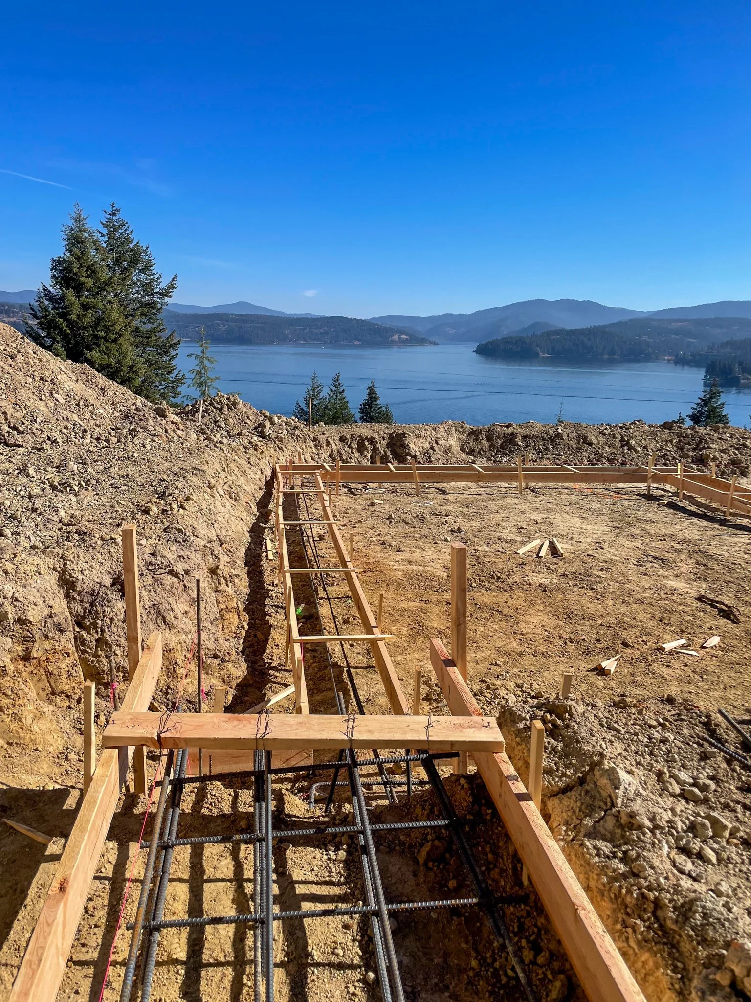 Construction site with wooden framing and steel rebar, overlooking a lake with trees and mountains in the background under a clear blue sky.