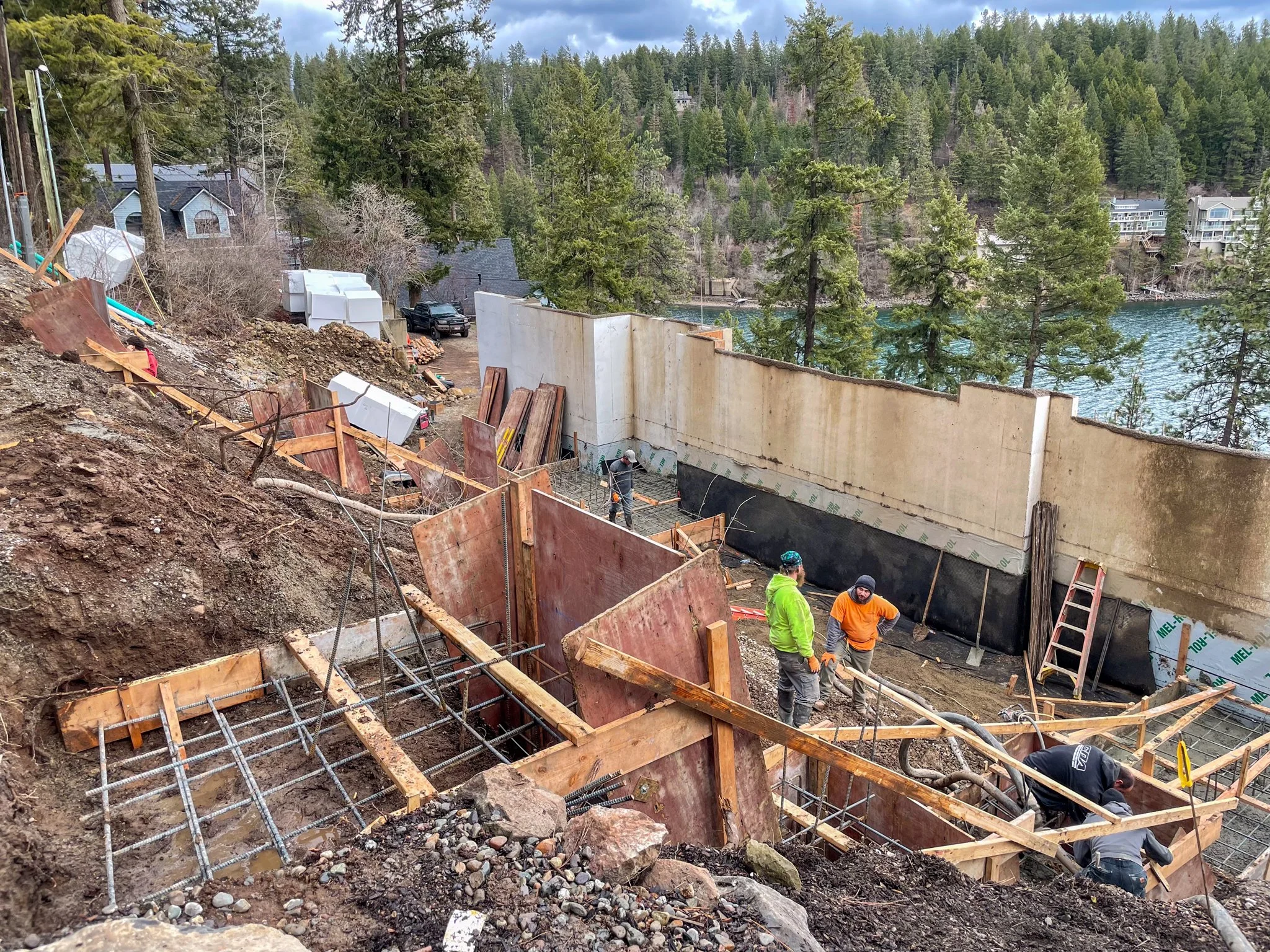 Construction site on a hillside near a lake, with workers and concrete wall framework, surrounded by tall trees and residential buildings in the background.