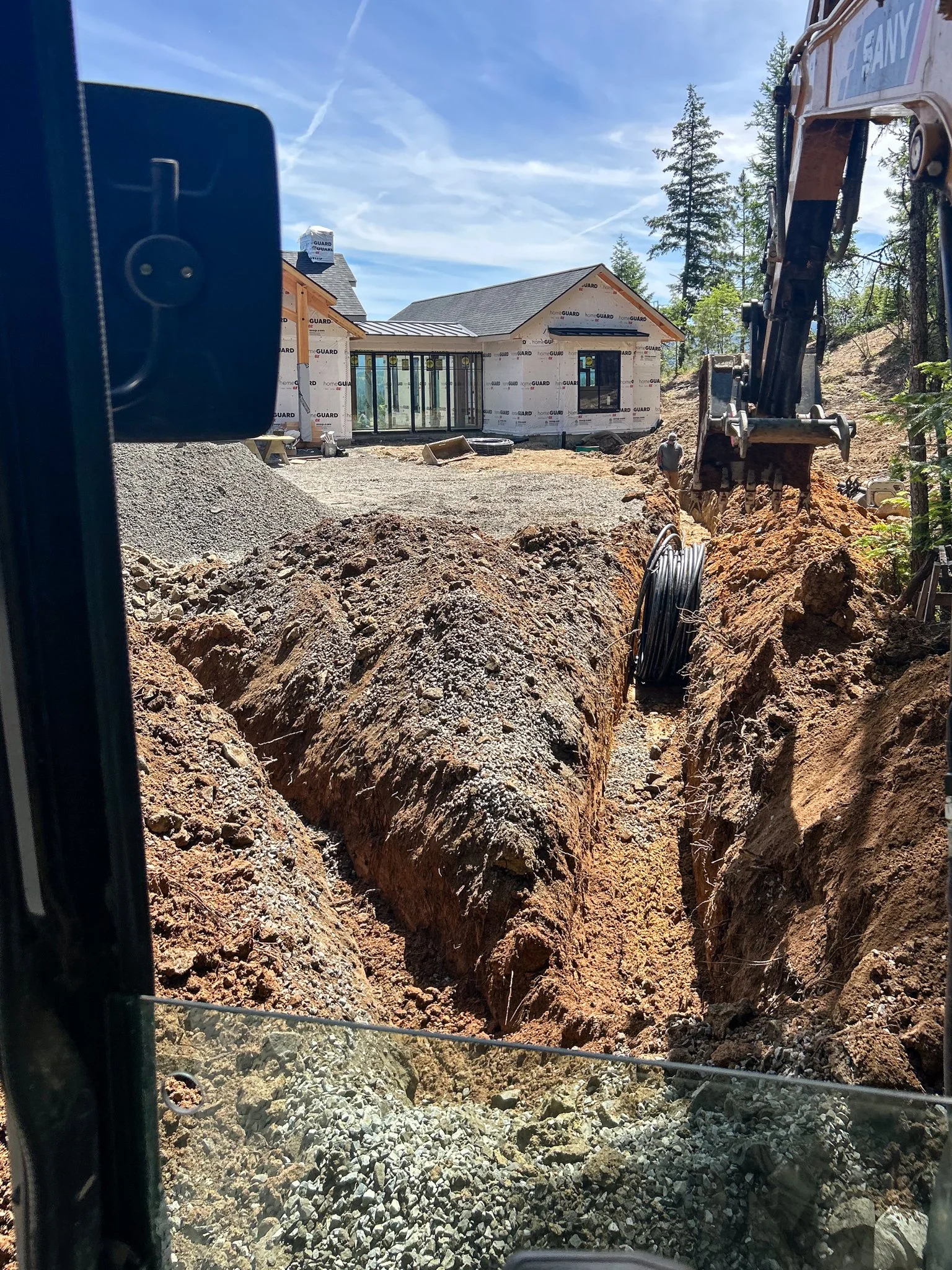 Construction site with a house under construction, excavator digging a trench, piles of dirt and gravel, trees in background, clear blue sky.