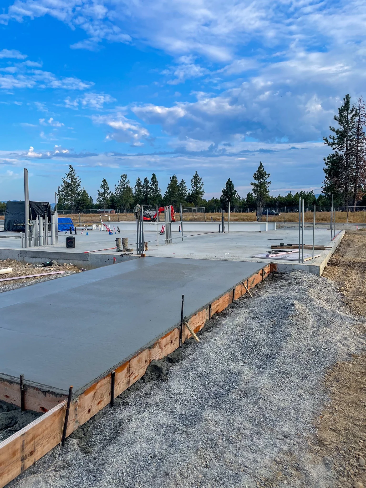 Construction site showing a concrete foundation with surrounding wooden formwork and construction materials, with trees and a partly cloudy sky in the background.
