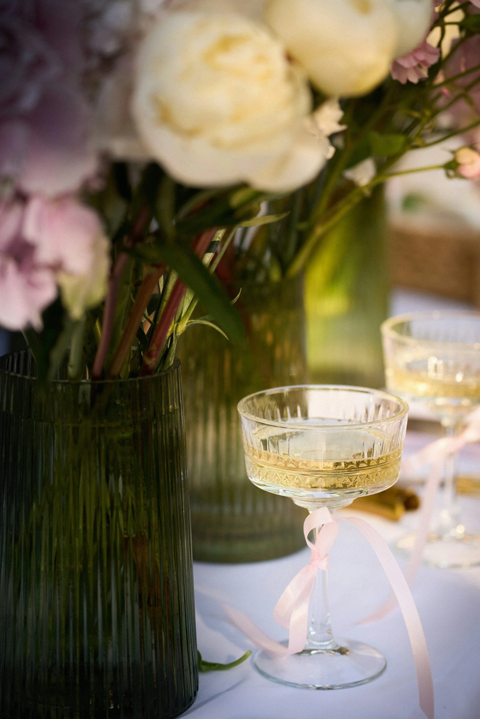 A floral arrangement in a dark green vase with white and pink flowers next to a glass of champagne with a pink ribbon tied around the stem.