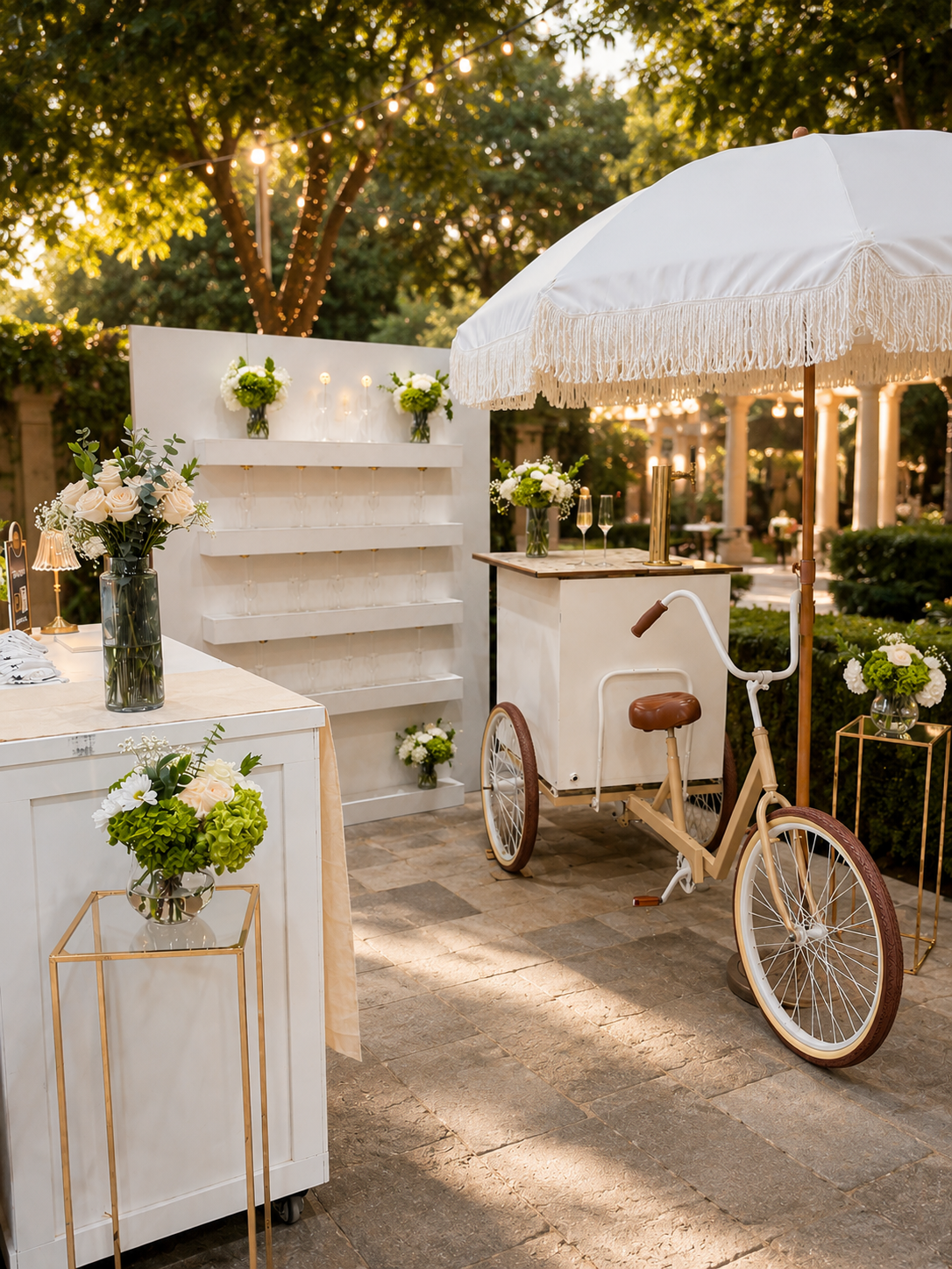Outdoor event setup with white flowers, a tricycle serving as a bar under a large white umbrella, and string lights in a garden.