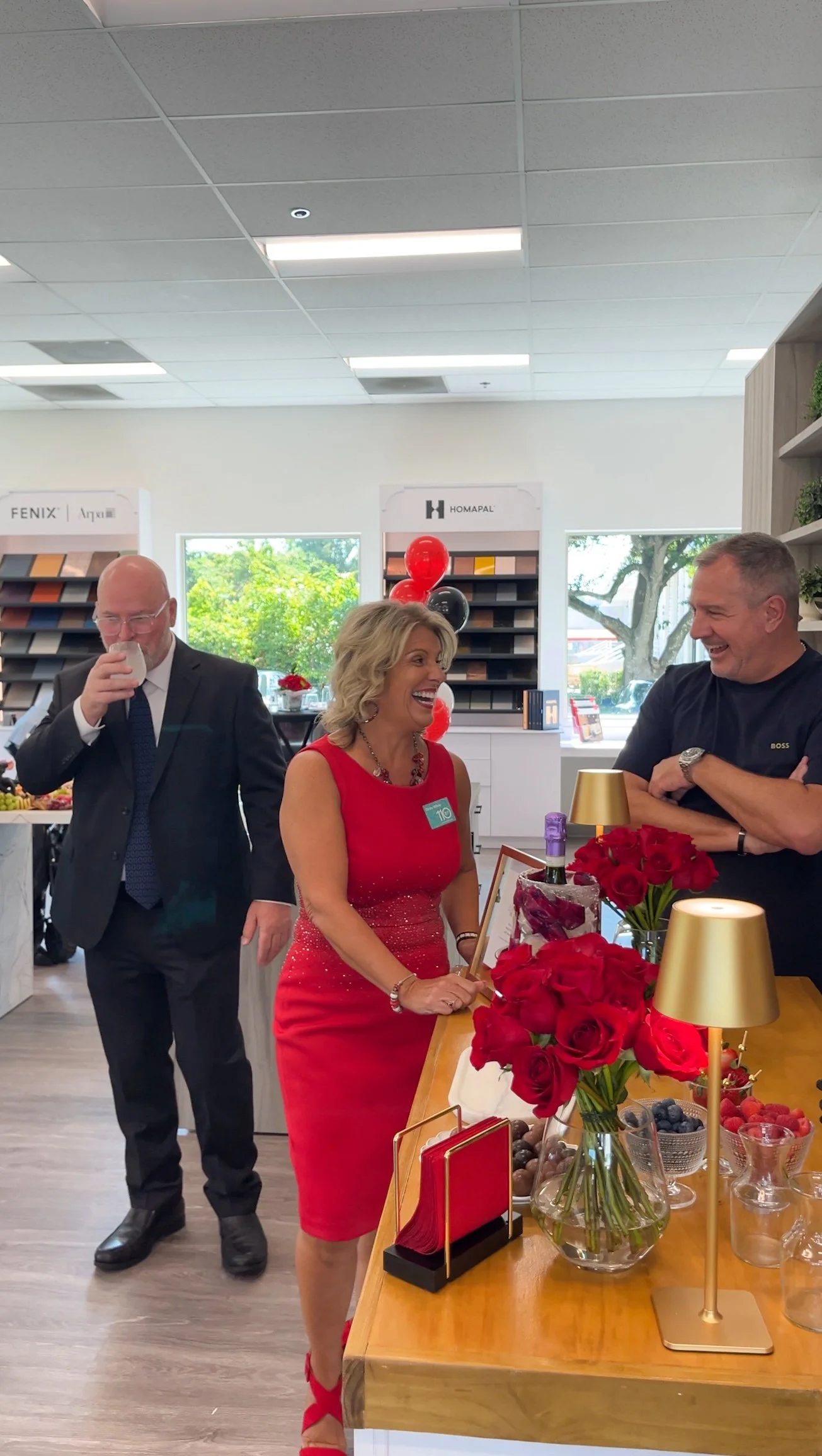 A woman in a red dress with a name tag is standing by a table filled with red roses, candles, and various desserts, smiling and conversing with a man in a black shirt. Two other people, a man in a suit and a woman not fully visible, are also present in the background.
