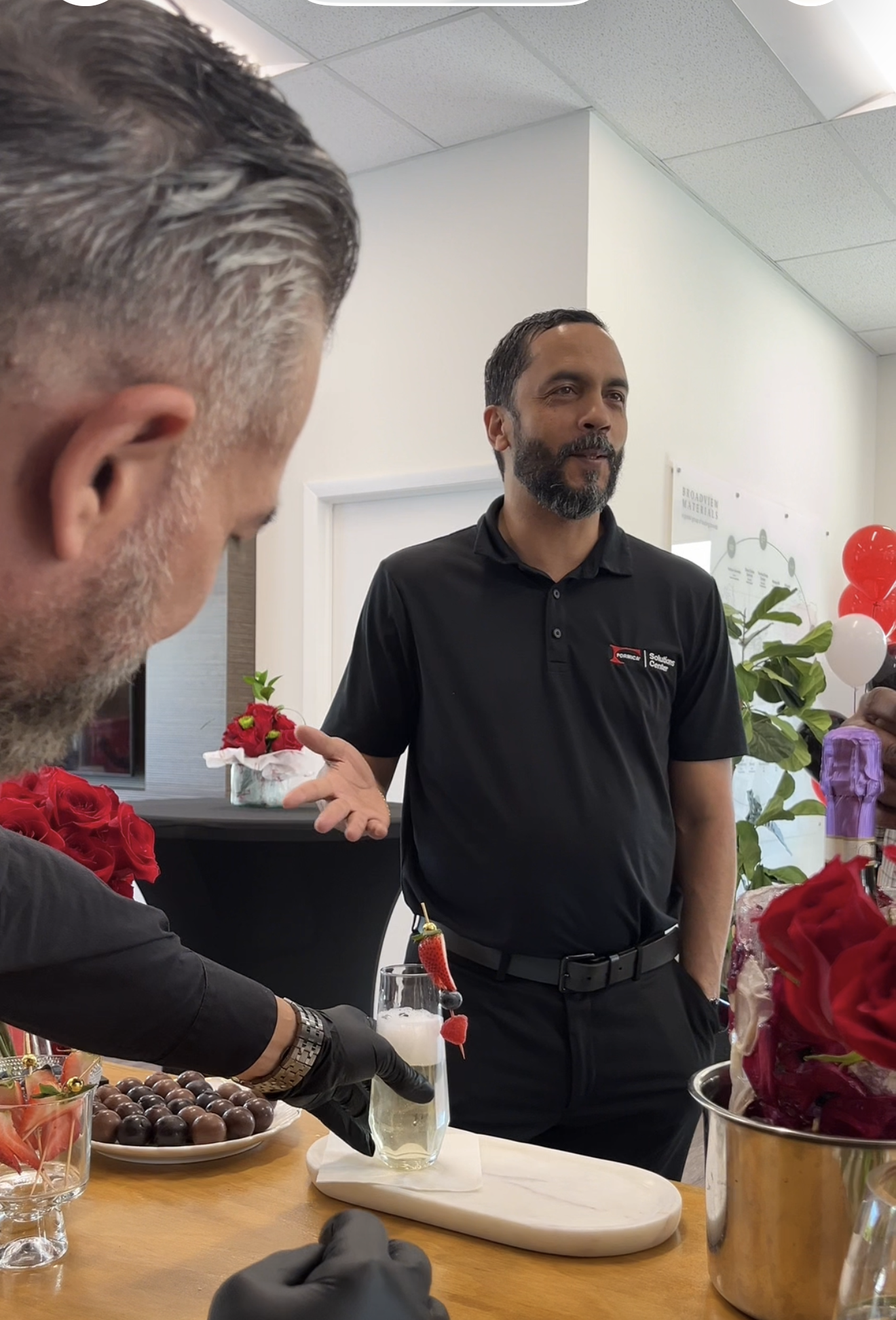 A man with a beard in a black polo shirt talking at a celebration table with desserts and flowers, and a person with gray hair and a beard serving chocolate candies.