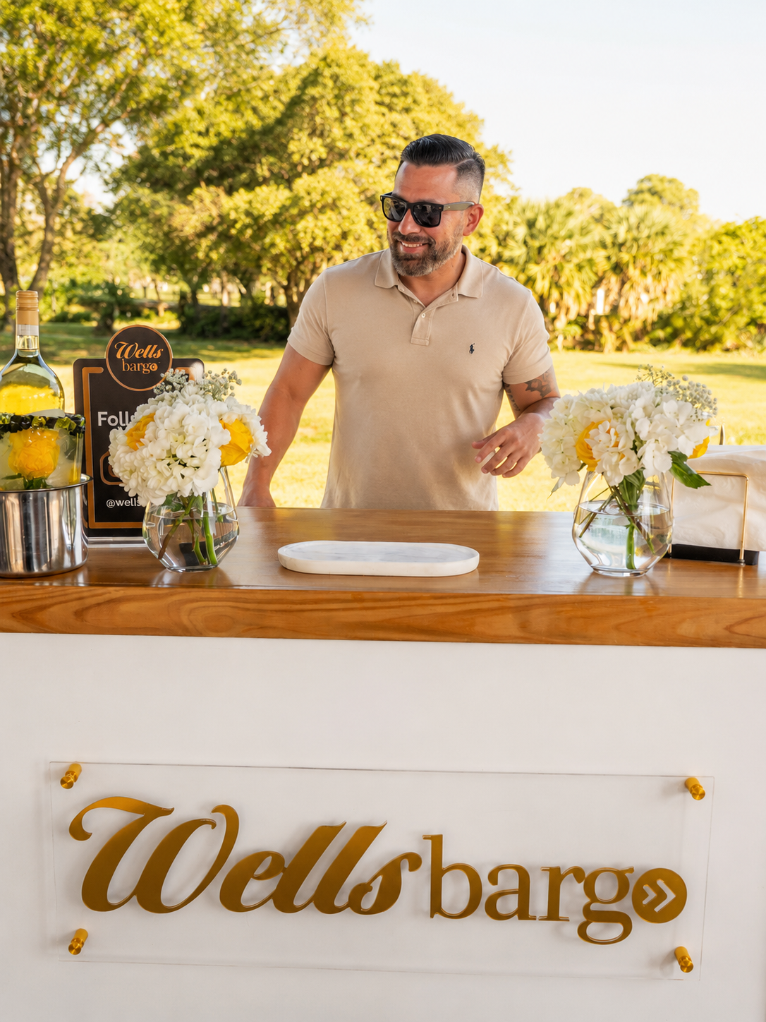 A man with dark hair, beard, sunglasses, wearing a beige polo shirt, stands behind a wooden bar outdoors, decorated with white and yellow floral arrangements. There are bottles on the left, and a sign with the Wells Bargo logo on the front of the bar.