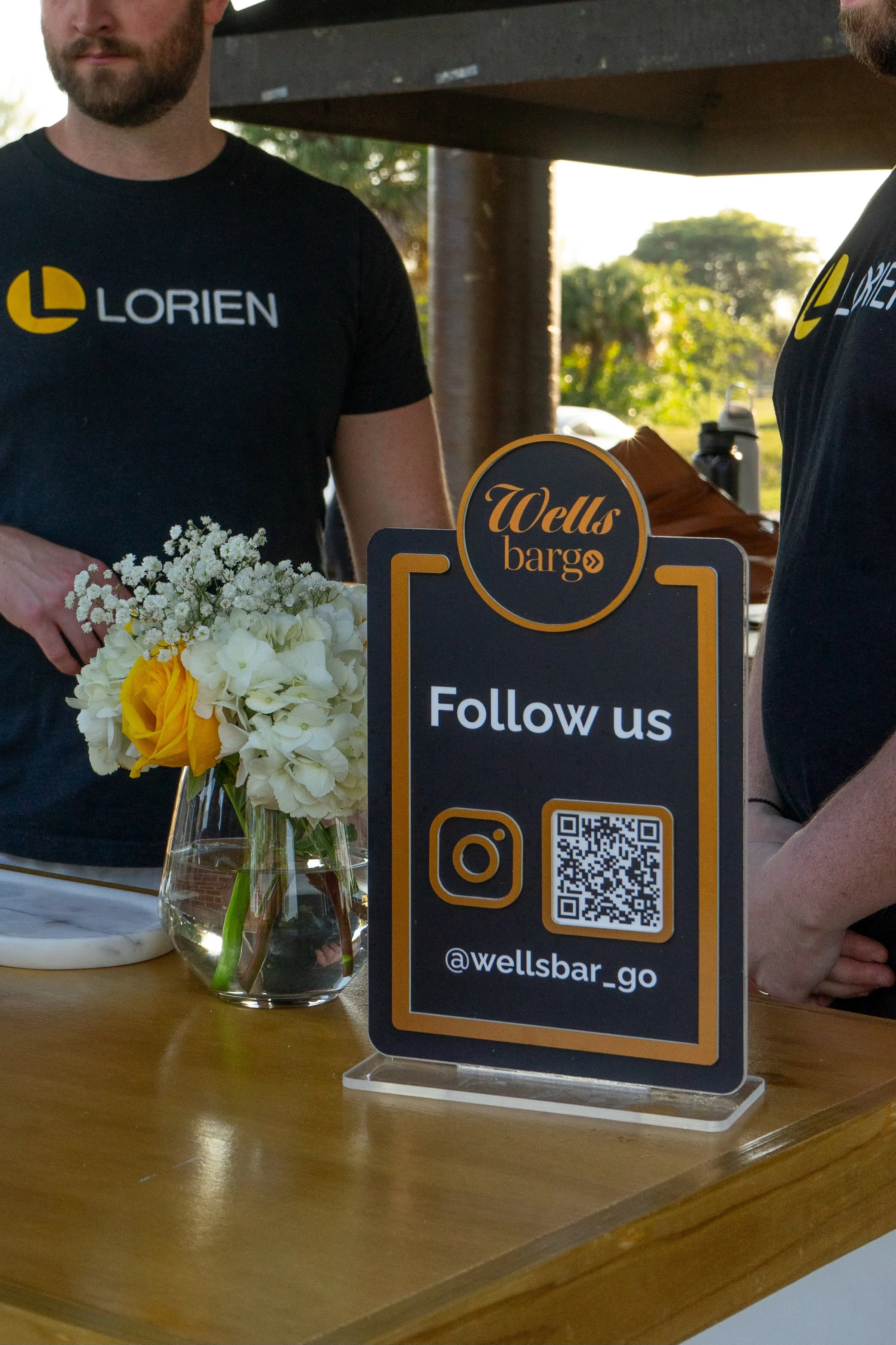 People wearing black T-shirts with the logo and text for 'Lorien' and 'Wells bar' standing at a table with a glass vase of flowers and a sign encouraging social media following, including a QR code and account handle '@wellsbar_go'.