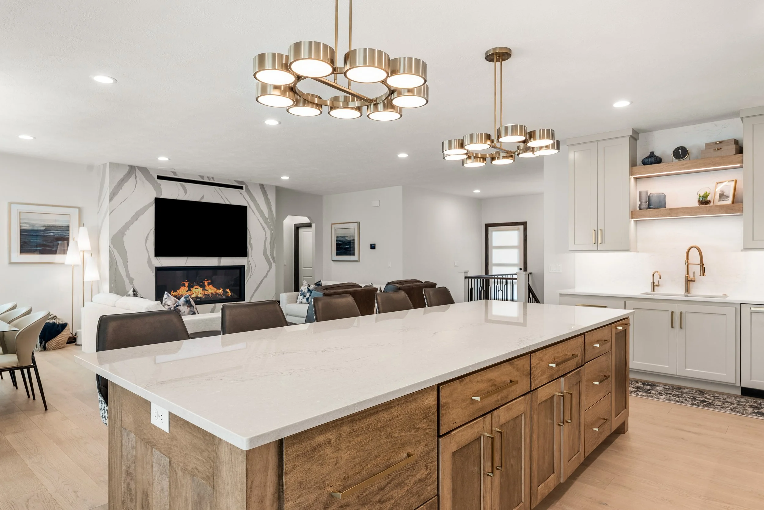 Modern kitchen with a large white island, wooden cabinets with gold handles, and two contemporary gold and silver chandeliers.