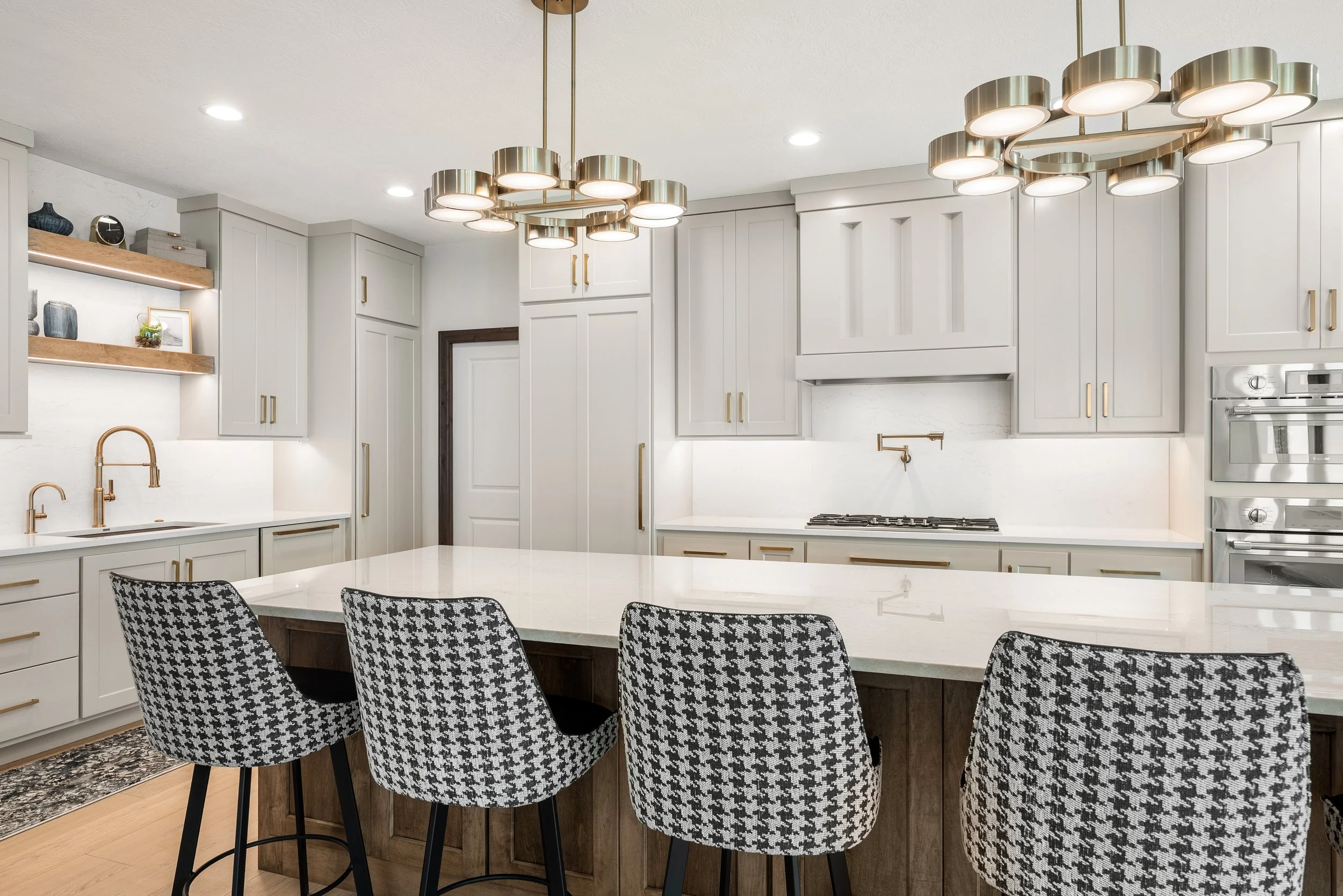 Modern kitchen with white cabinets, gold handles, a white marble backsplash, and a large white island with a marble countertop. Hanging statement chandeliers with multiple gold and white lights; patterned black and white chairs surround the island. Open shelving with decorative items, and built-in double ovens visible.