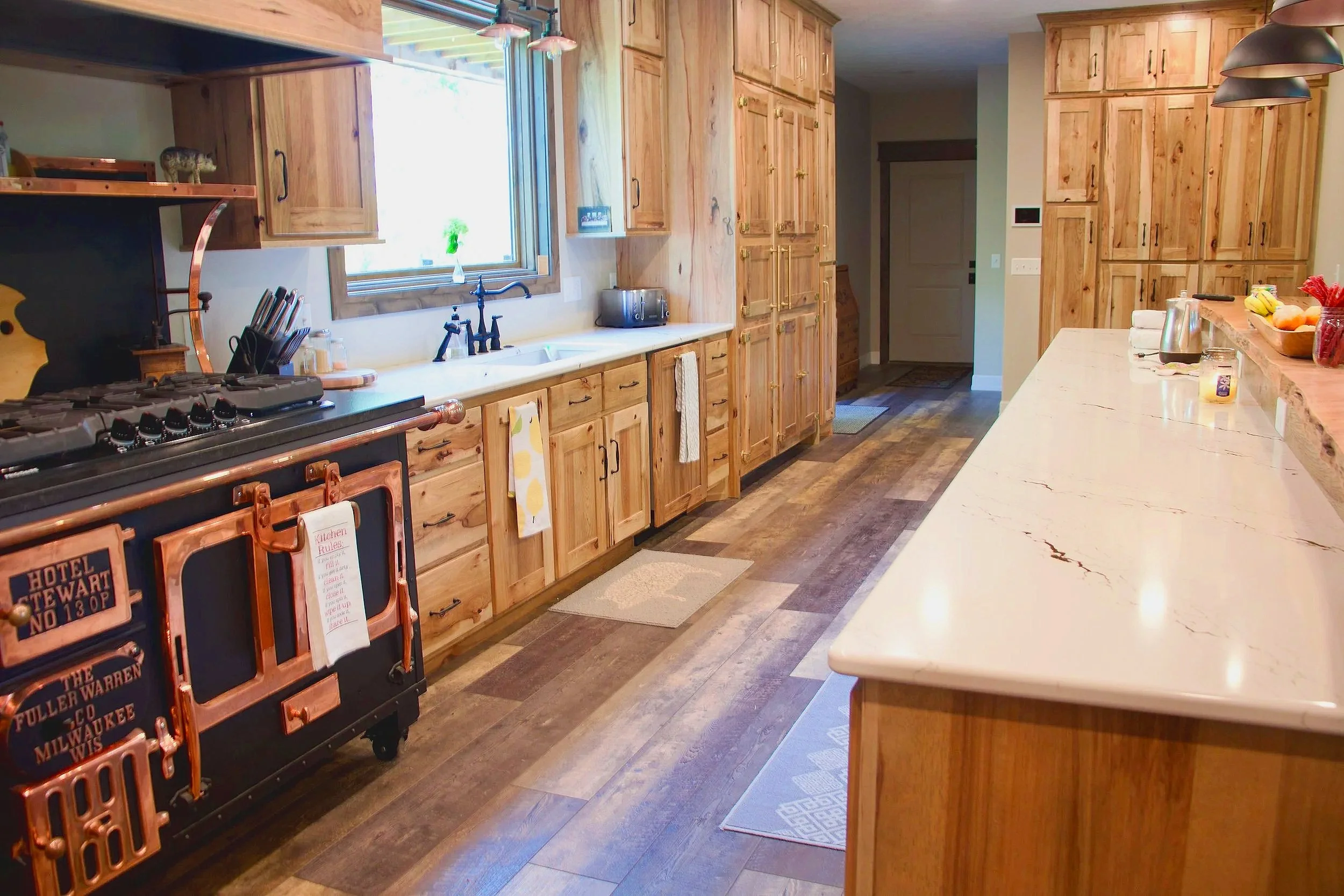 A kitchen with wooden cabinets, a marble countertop, and a window above the sink. There is a stove on the left, some kitchen utensils, a toaster, and a small candle on the counter. The kitchen has a warm, rustic style with wood-paneled floors.