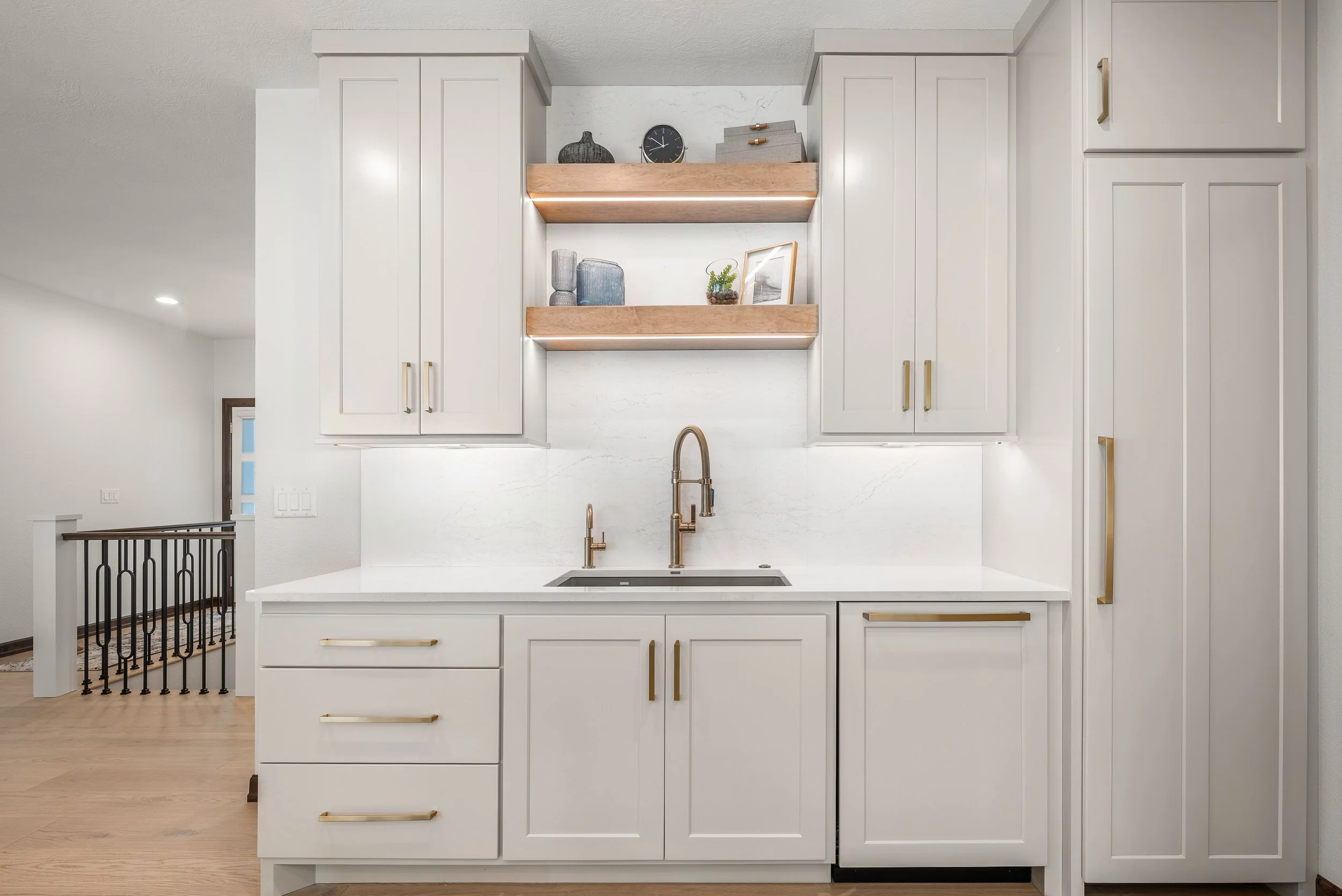 Modern white kitchen with gold hardware, a gold faucet, open wooden shelves with decor, and a black clock on the marble backsplash.