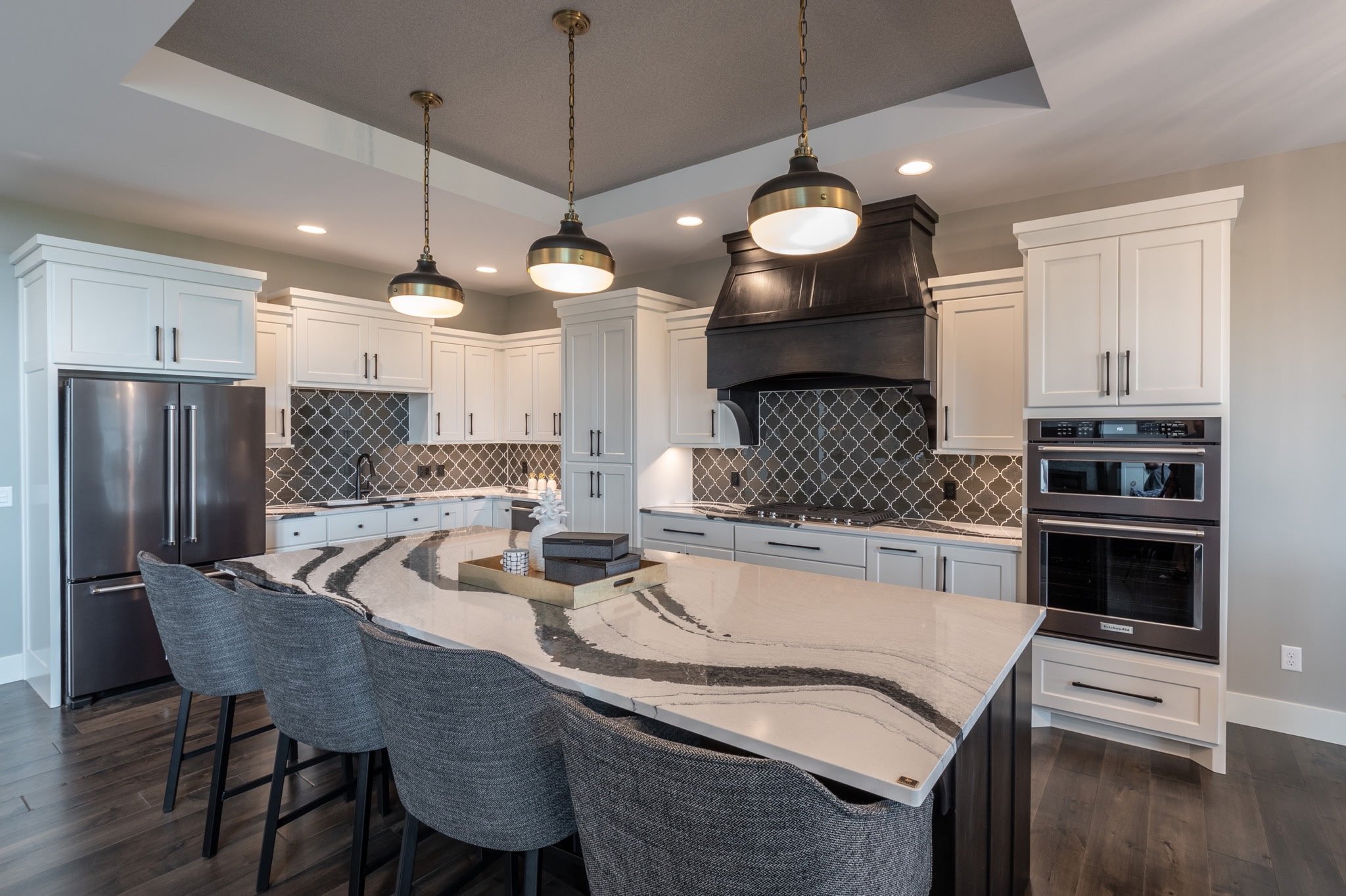 Modern kitchen with white cabinets, black hardware, black and gray patterned backsplash, stainless steel refrigerator, wall oven, black range hood, and a large island with a marble countertop and four gray upholstered chairs.